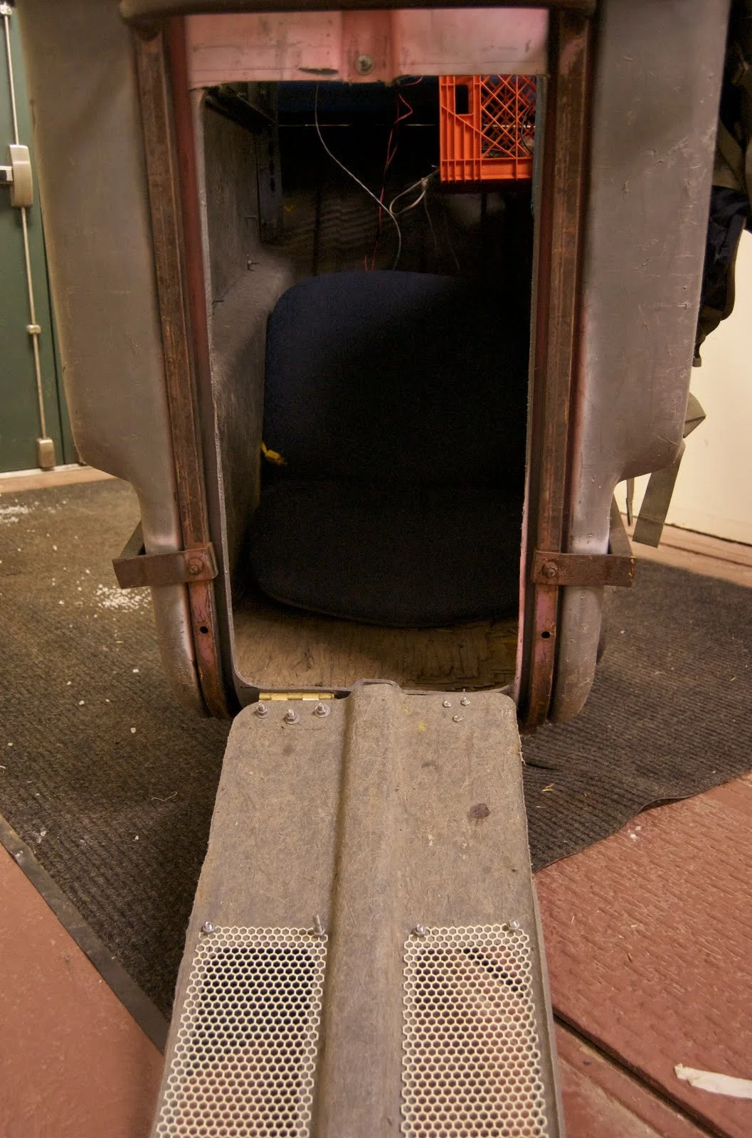 Inside an old, metal robot costume with a black fabric interior seat and a small ramp at the opening, showing exposed wires and a plastic crate in the background.