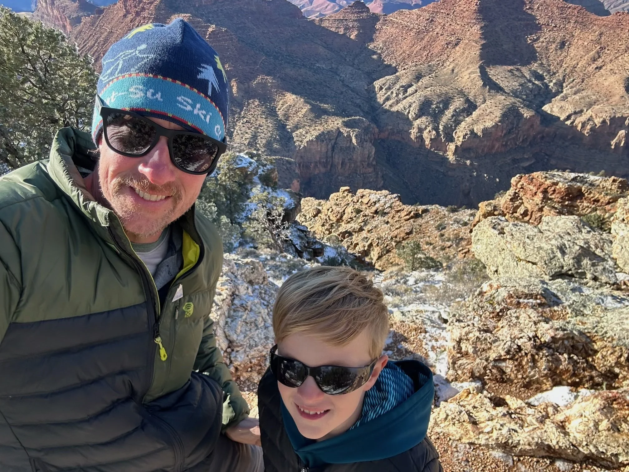 A man wearing sunglasses and a red head covering outdoors with a child on his back, both dressed for cold weather, with bare trees and a clear blue sky in the background.