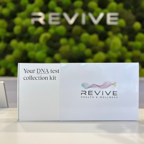 A DNA test collection kit from REVIVE Health & Wellness on a white surface with a green leafy background and the word REVIVE spelled out in large white letters on a green hedge.