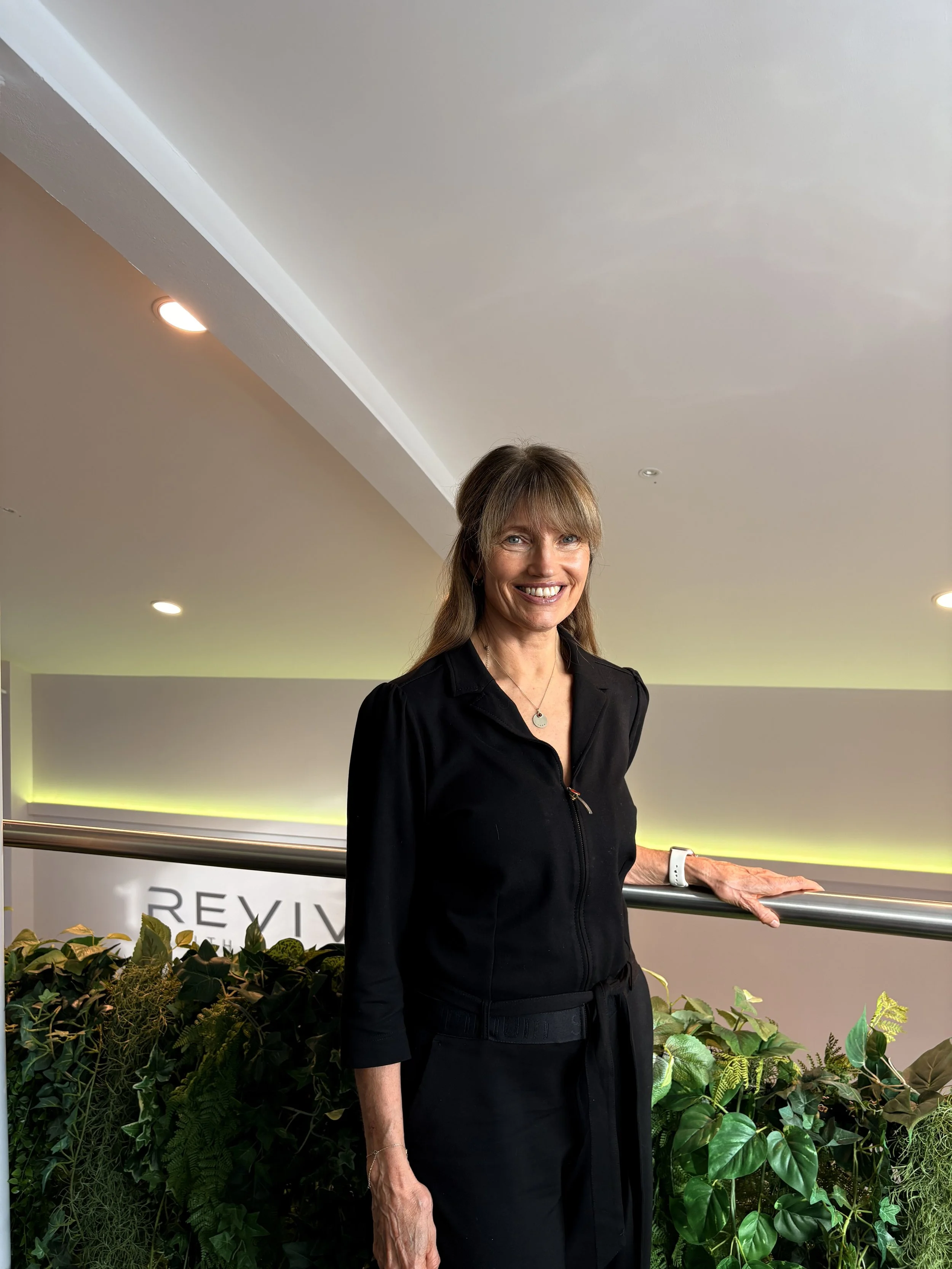 A woman with brown hair styled with bangs, wearing a black outfit, smiling and standing near a railing with green plants behind her.