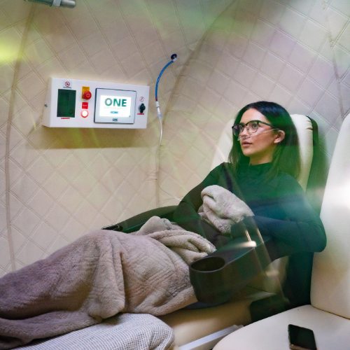 A woman sitting in a medical treatment chair with a blanket, wearing glasses, holding a mug, in a clinical room with medical devices on the wall.