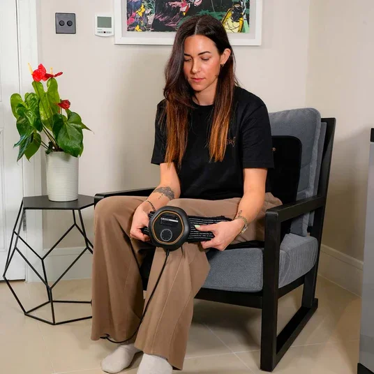 A young woman with long brown hair sitting on a gray chair, holding a portable speaker in her lap, in a room with a white wall, colorful artwork, a side table with a potted plant, and beige walls.
