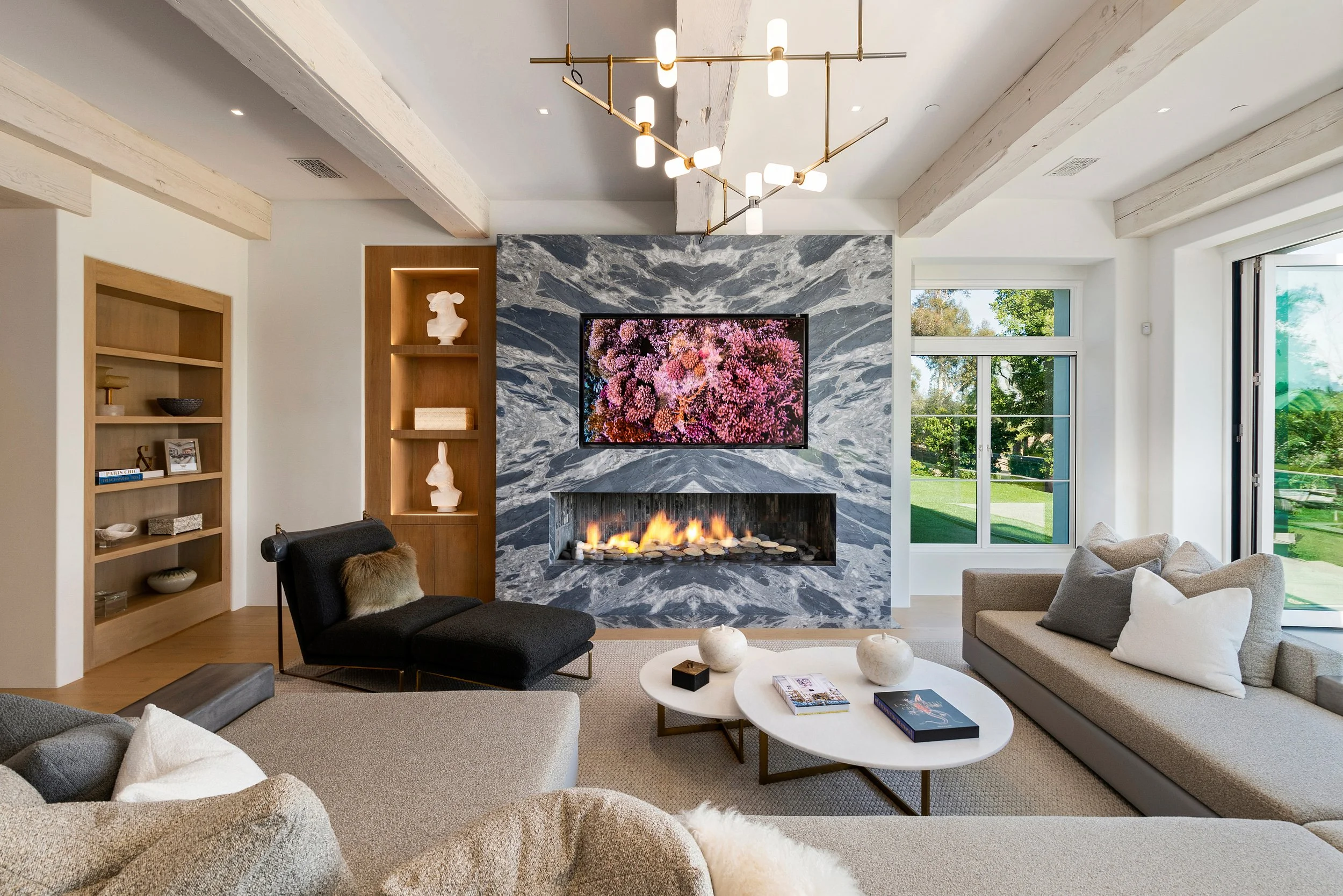 Modern living room with a marble fireplace, flat-screen TV, and large windows showing greenery outside. The room features a contemporary chandelier, built-in shelves with decorative items, and neutral-colored furniture.