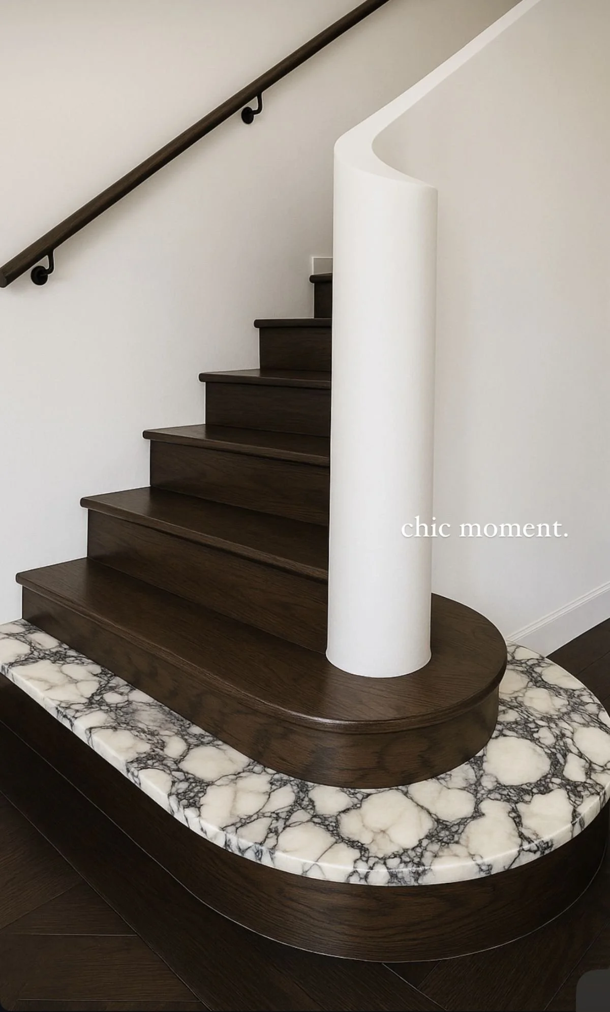 A modern staircase with dark wood steps and a white curved wall at the side, positioned above a marble-topped piece of furniture. The words 'chic moment.' are written on the wall.