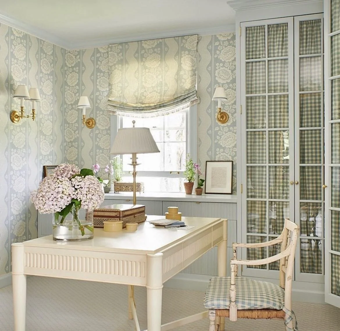 A bright, cozy room with floral and checkered wallpaper, a white desk with a vase of pink hydrangeas, a lamp, potted plants, and framed picture, a checkered upholstered chair, and a large window with a floral window treatment.