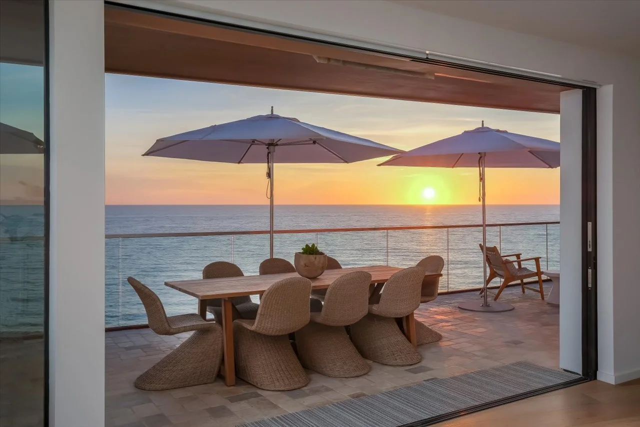 Oceanfront balcony with a wooden dining table, wicker chairs, two umbrellas, and a sunset view over the sea.