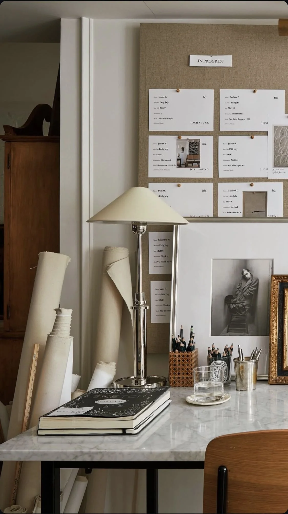 An interior workspace with rolled-up paper, books, and drawing supplies on a marble table, a lamp, and a wall with various notes, artwork, and a black-and-white photograph of a person in the background.