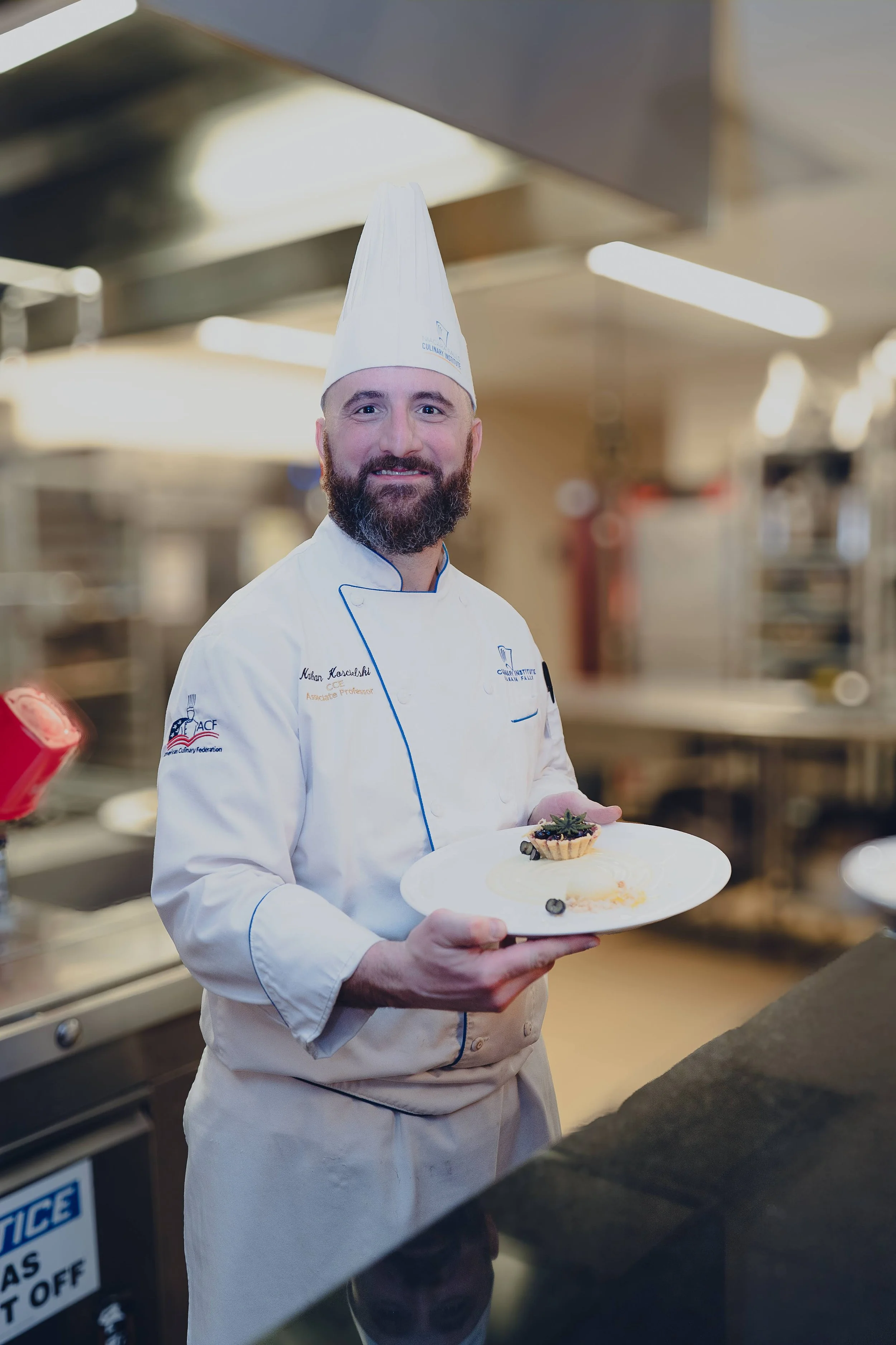 Chef smiling in a commercial kitchen holding a white plate with a small tart and a spoon of dessert garnished with blueberries.
