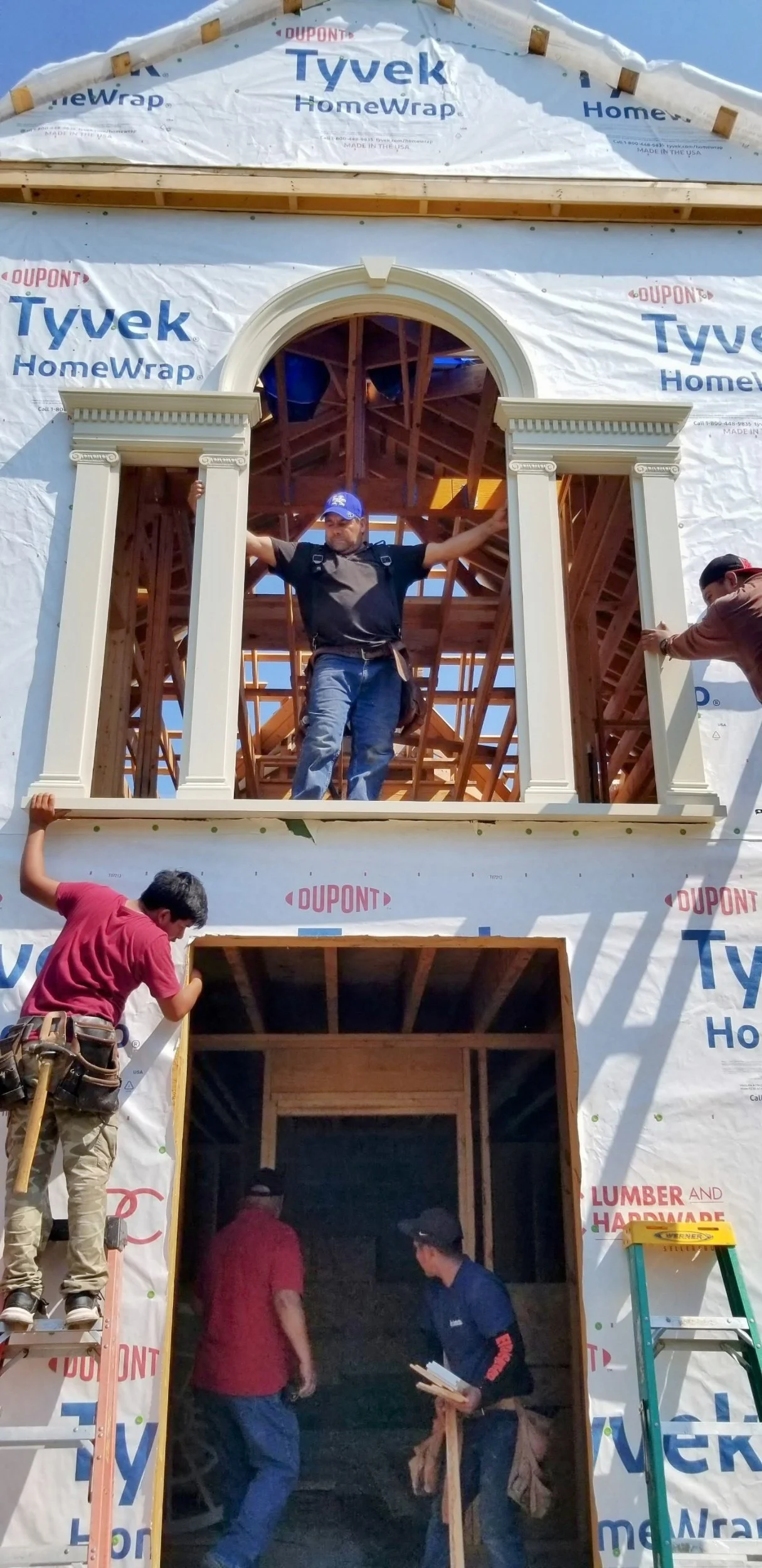 Construction workers installing a decorative arched window frame on the exterior of a building under construction, with house wrap labeled 'Tyvek HomeWrap' and scaffolding around.