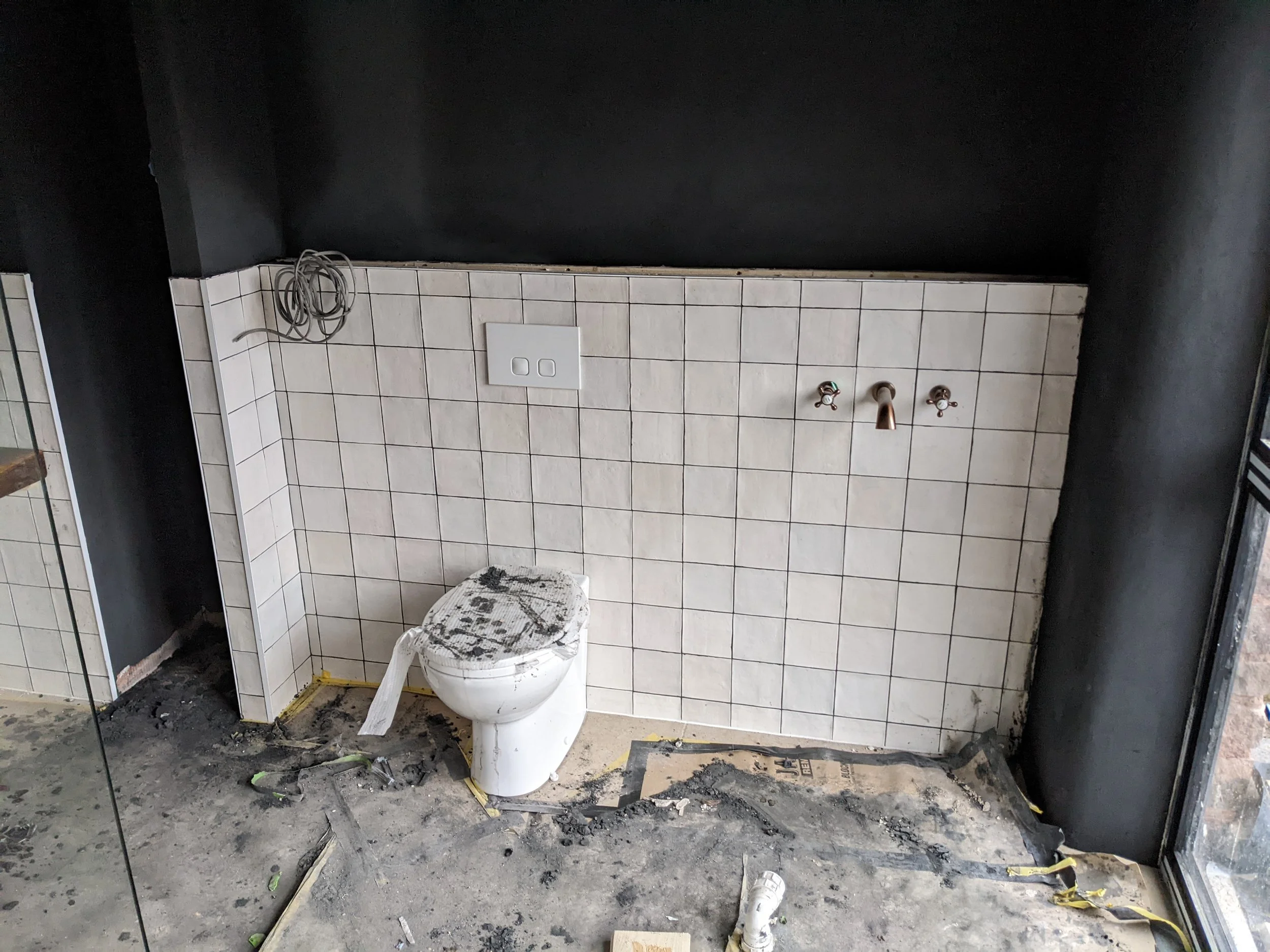 Qube Bathrooms - Progress photo of previous Bathroom Installation. A room under construction with beige tile wall that has plumbing fixtures, a black wall above, and dirty concrete floor.
