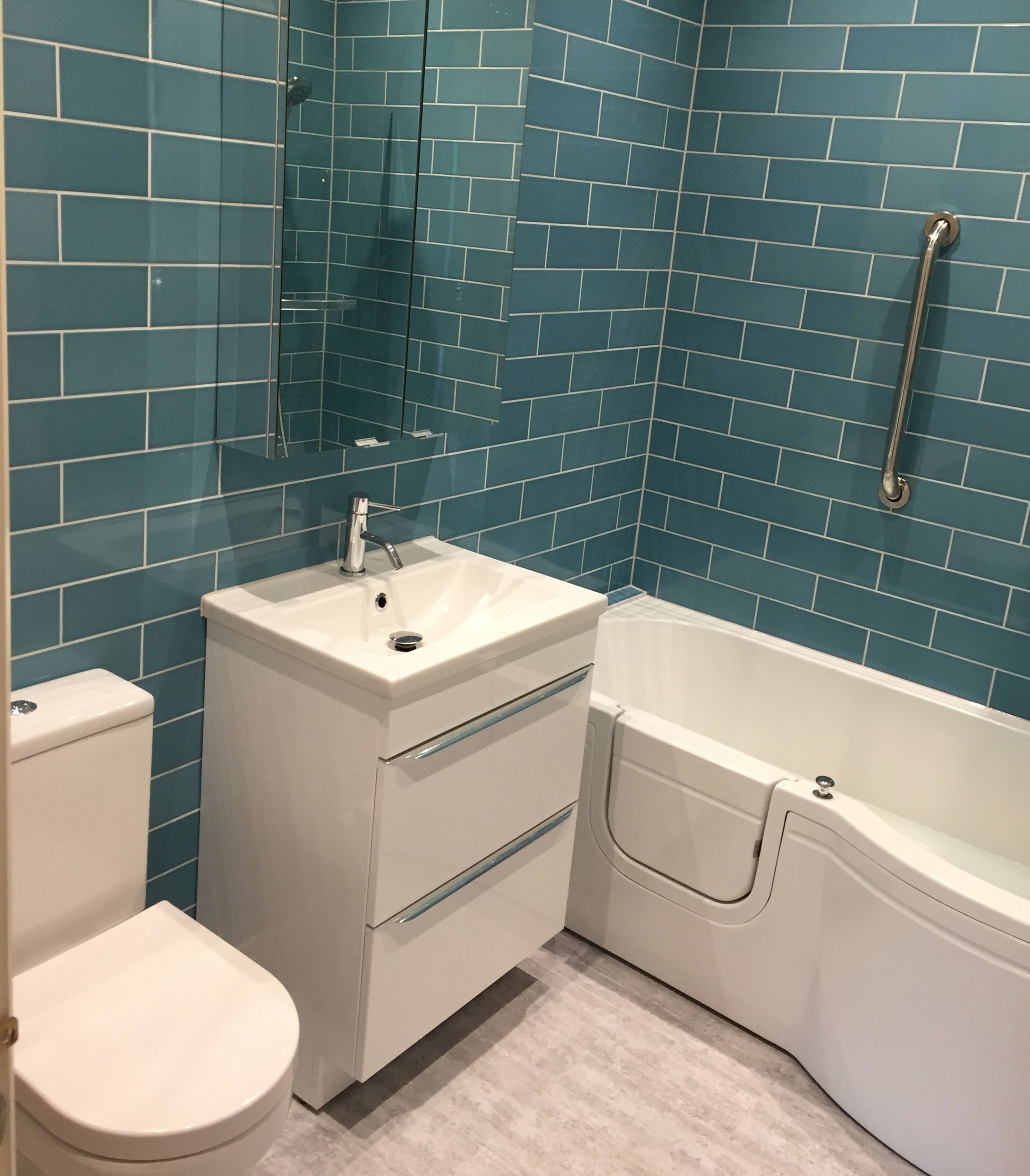 Qube Bathrooms, Bristol. Bathroom with blue tiled walls, white toilet, white vanity with sink, mirror, bathtub with a grab bar.