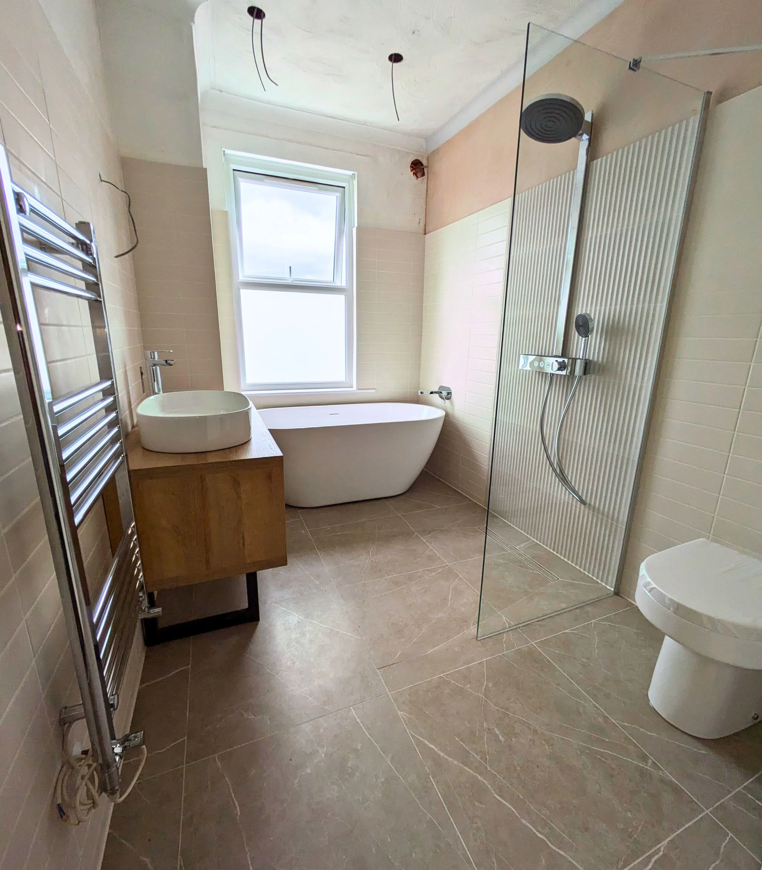 Qube Bathrooms. Wet room Installation. Modern bathroom with a freestanding bathtub, a walk-in shower with a glass partition, a wooden vanity with vessel sink, a window, and a wall-mounted towel radiator.