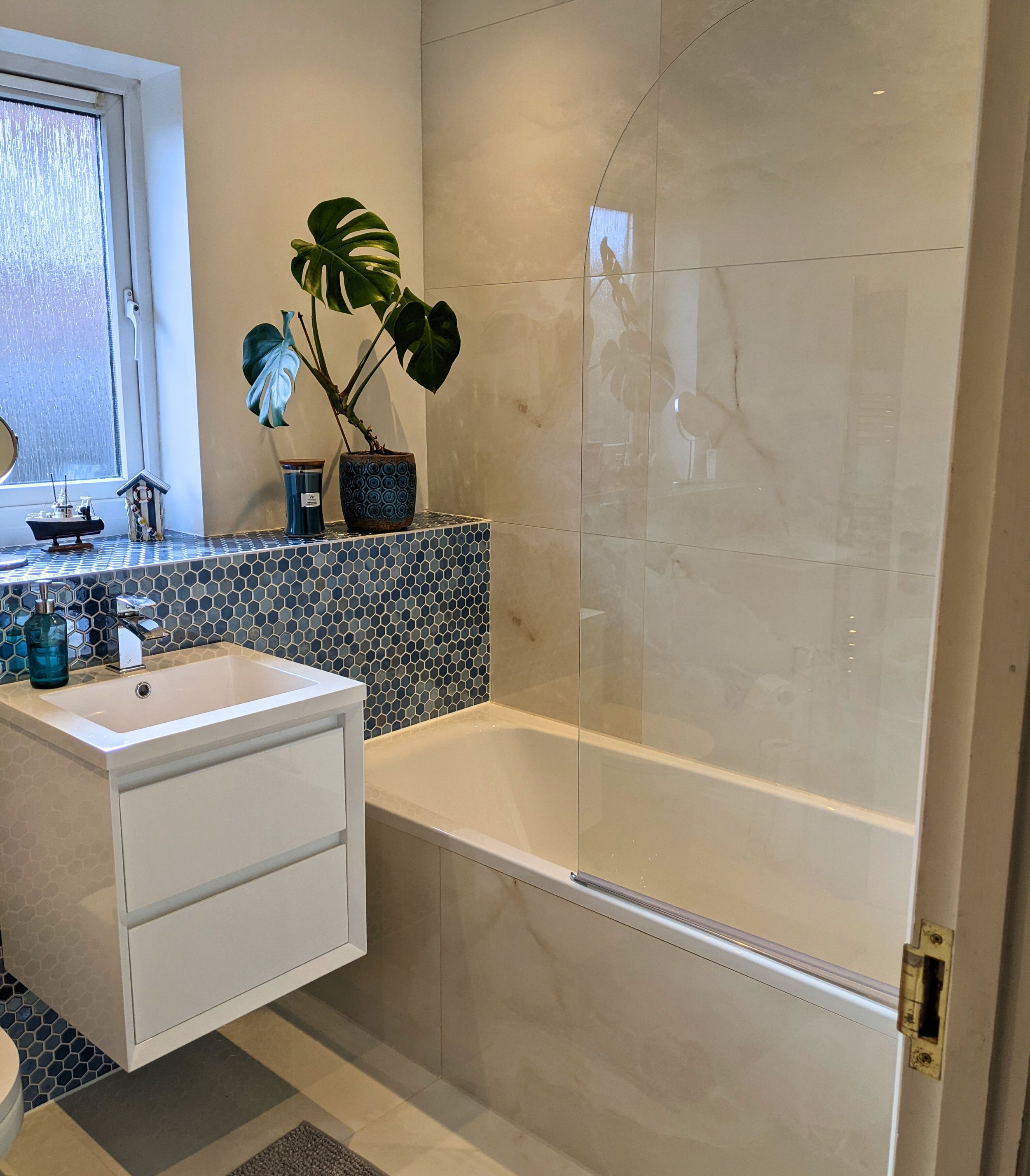 Qube Bathrooms - Photo of previous Bathroom Installation.Bathroom with a bathtub, a small white vanity with a sink, a window with daylight, blue mosaic tiles around the window ledge, a potted monstera plant, and various decorative items on the ledge.
