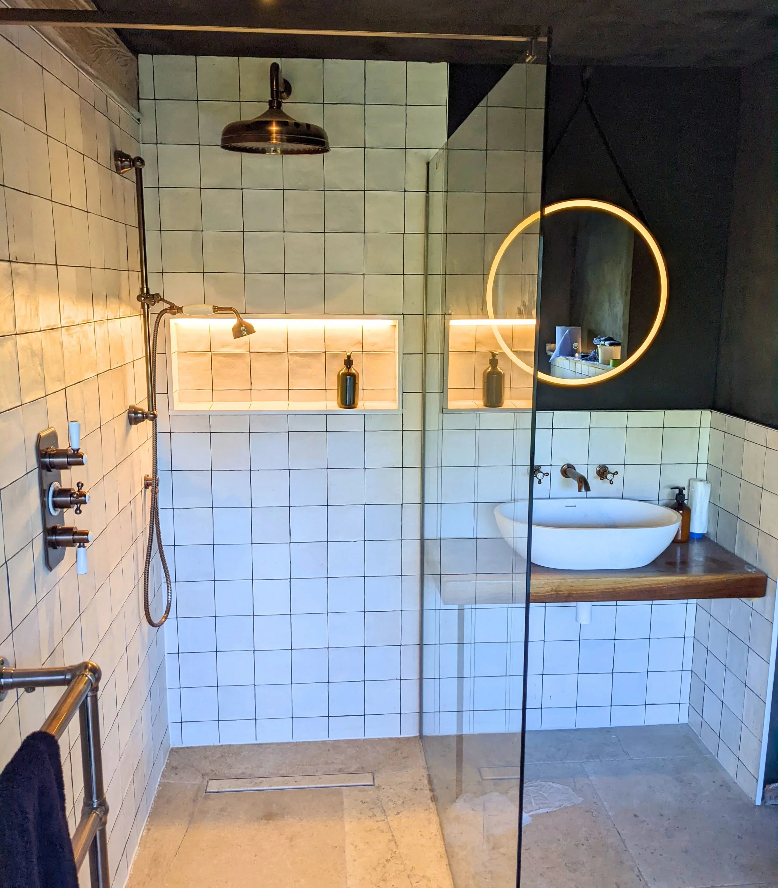 Qube Bathrooms Bristol. Modern bathroom featuring a walk-in shower with a rainfall showerhead, a built-in tiled shelf with a bottle, a circular illuminated mirror, a small sink with a wooden countertop, and a glass partition.