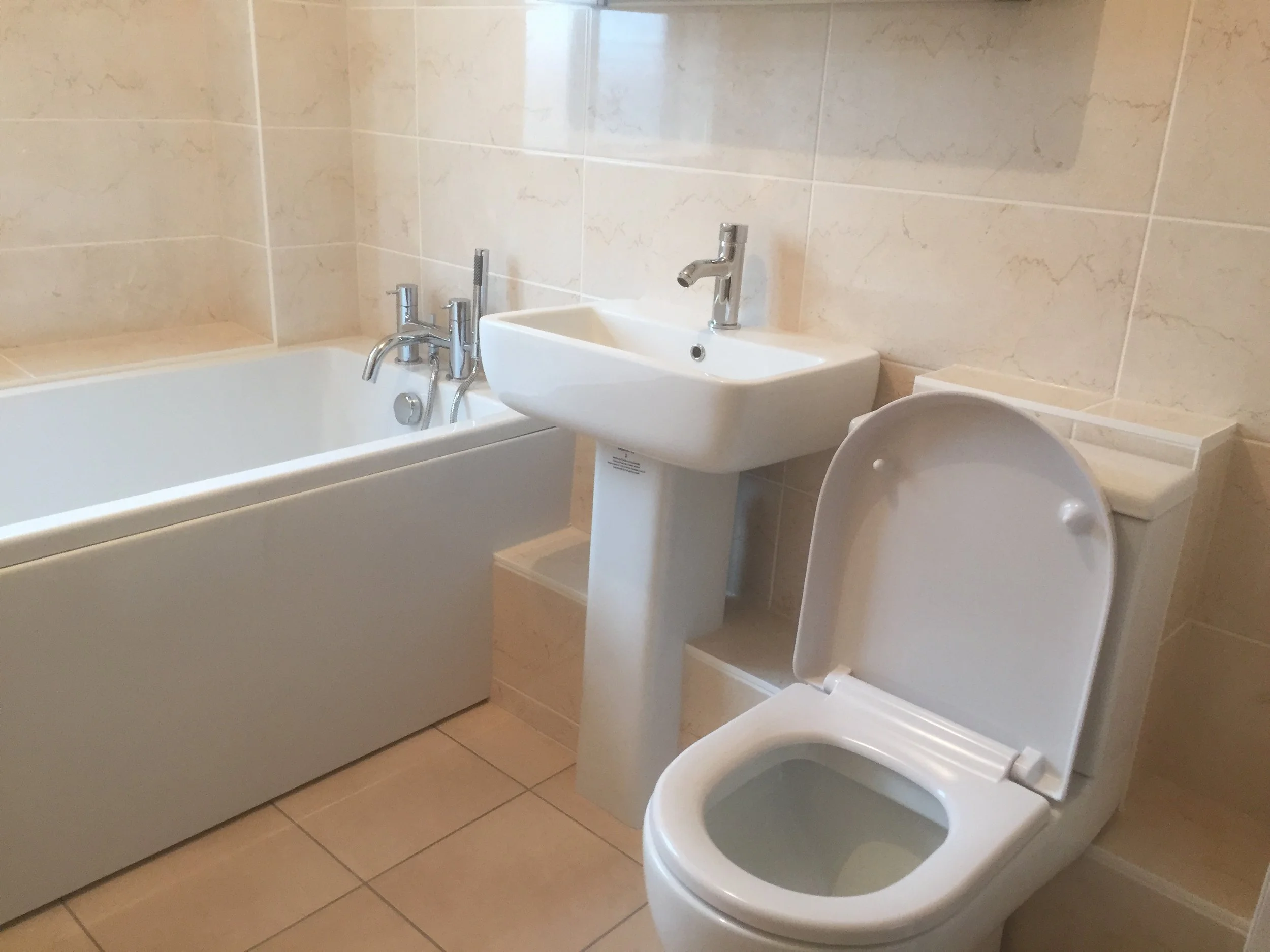 Qube Bathrooms, Bristol.  A bathroom featuring a bathtub with wall-mounted faucet handles, a small white sink with a single modern faucet, and a toilet with the lid open, all against beige tiled walls and floor.