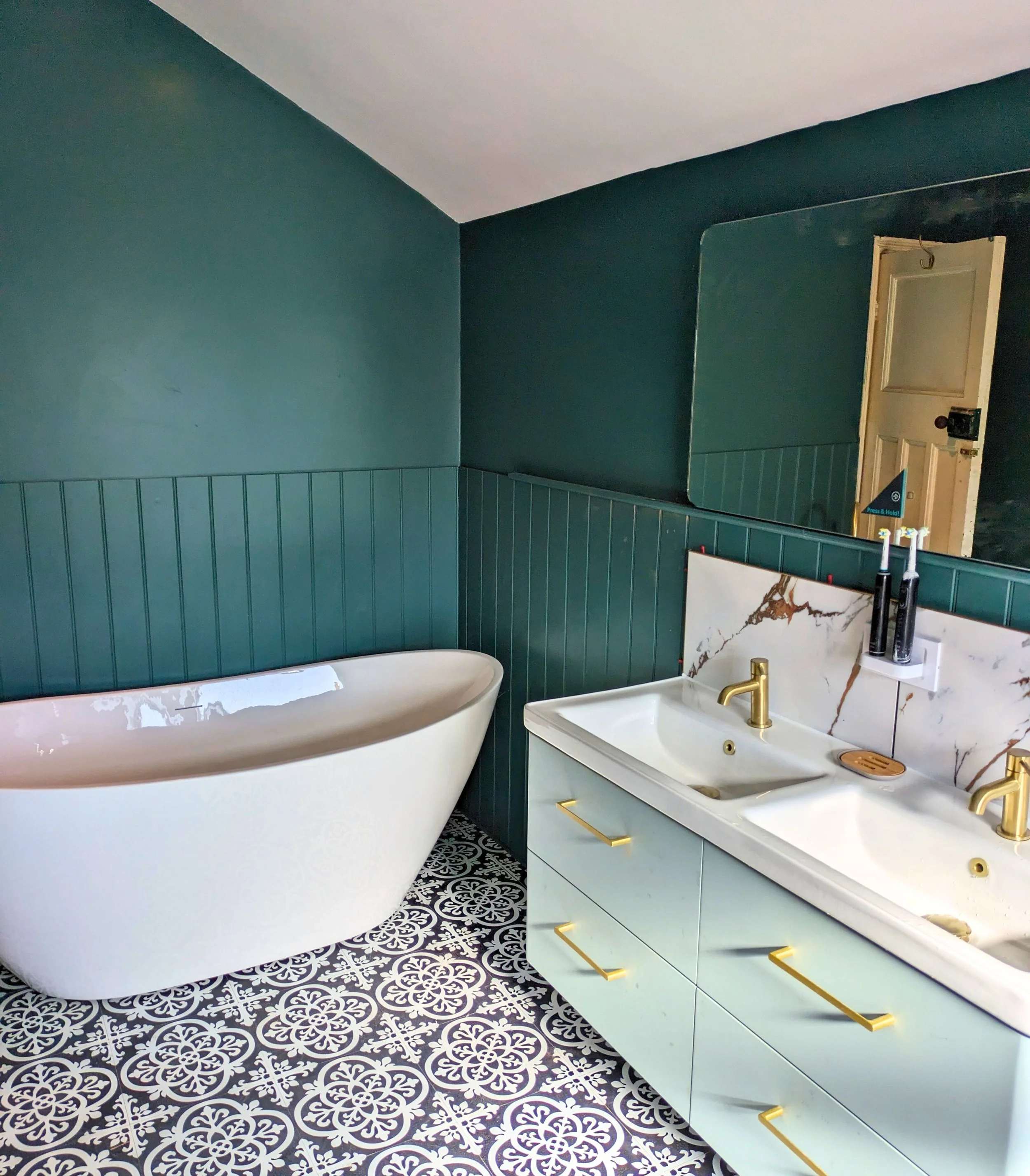 Qube Bathrooms - Photo of previous Bathroom Installation. Bathroom with a white freestanding bathtub, a double sink vanity with gold fixtures, and a large mirror on the teal-colored wall. The floor has black and white patterned tiles.