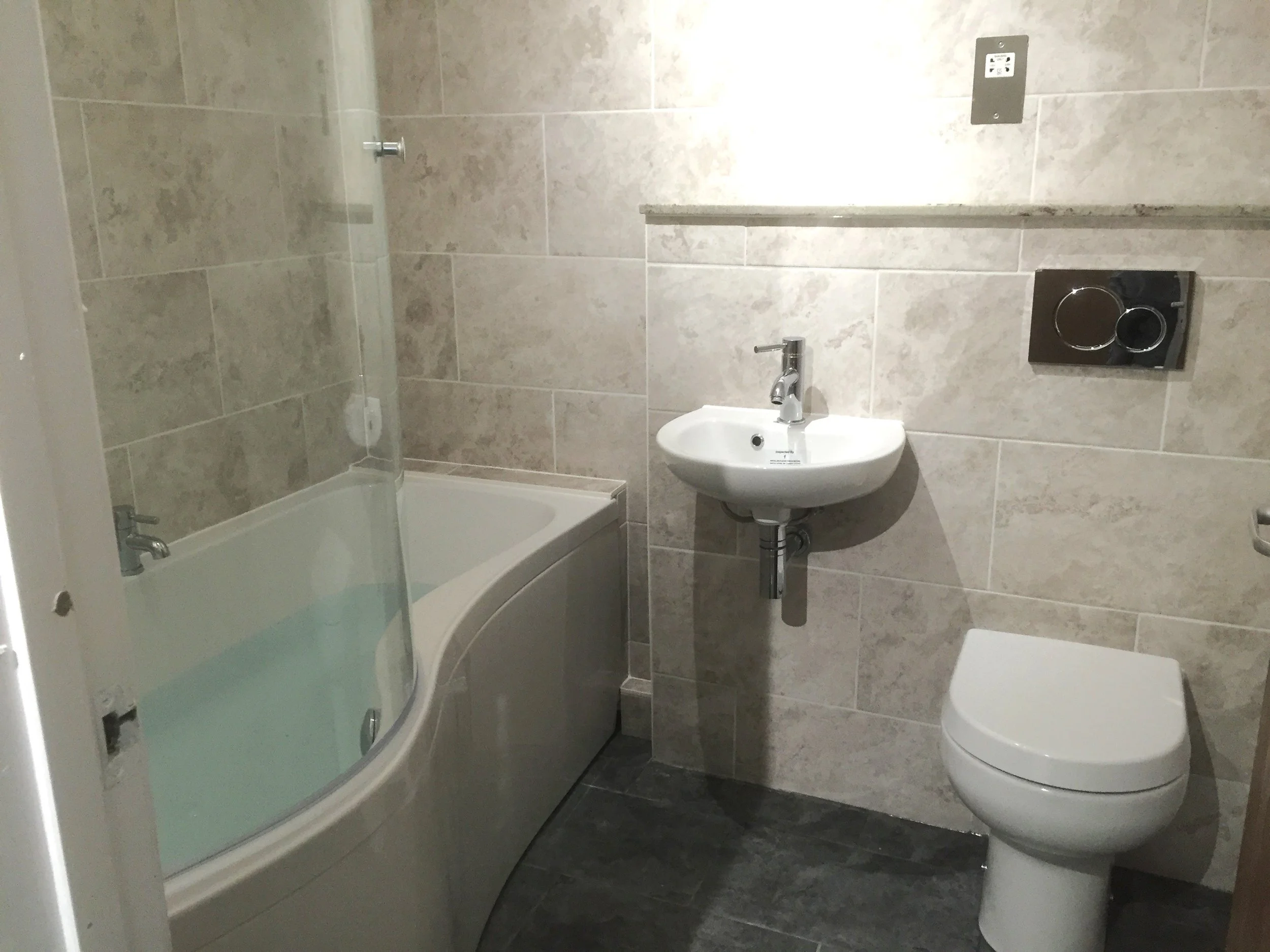 Qube Bathrooms, Bristol.  A small bathroom with beige tiled walls, a corner bathtub with a glass shower screen, a wall-mounted sink, and a wall-mounted toilet with a concealed cistern.