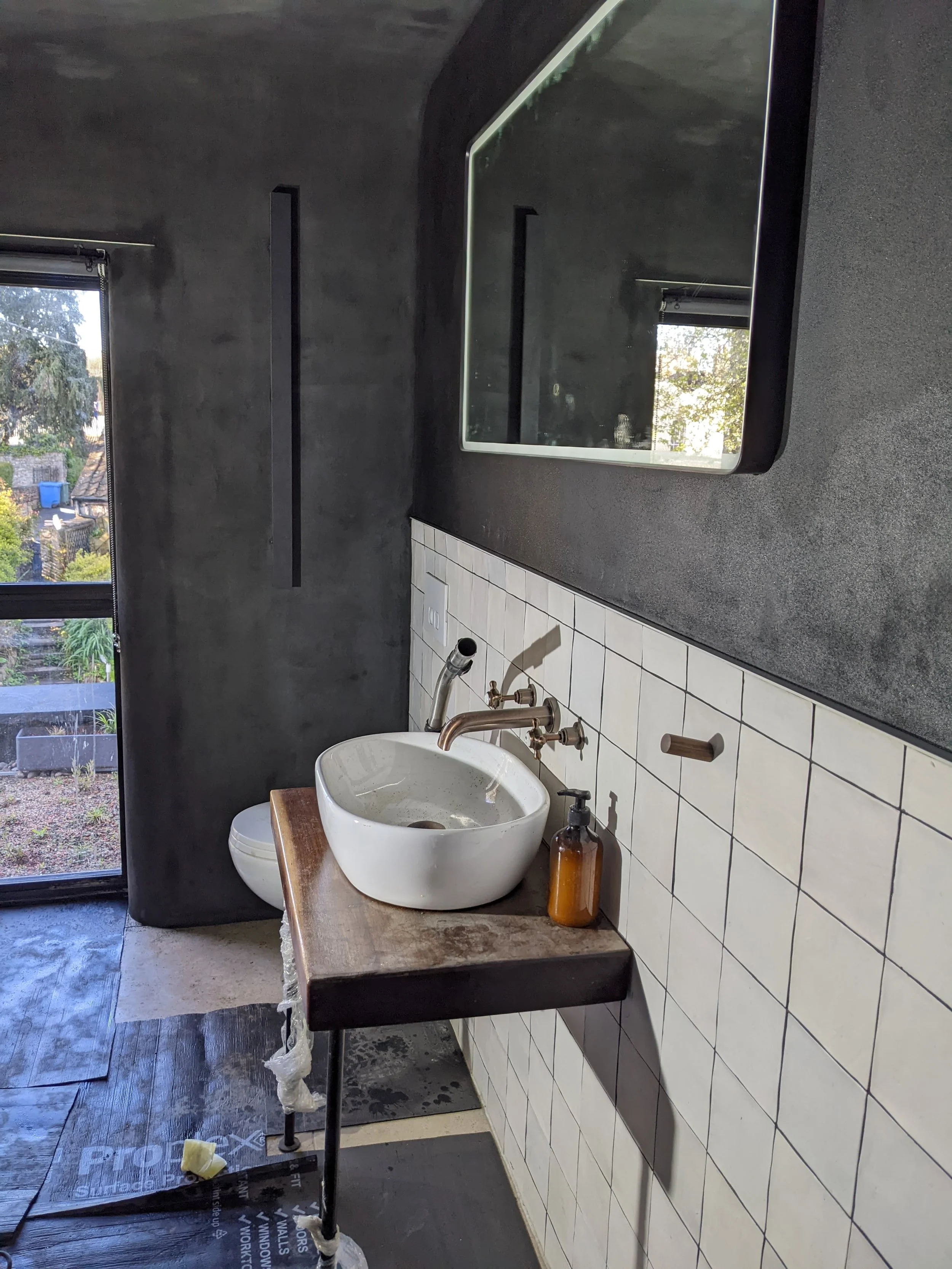 Qube Bathrooms, Bristol. Modern bathroom with a white vessel sink on a wooden countertop, a brown soap dispenser, dark wall and ceiling, a large mirror, and a window showing outdoor greenery.