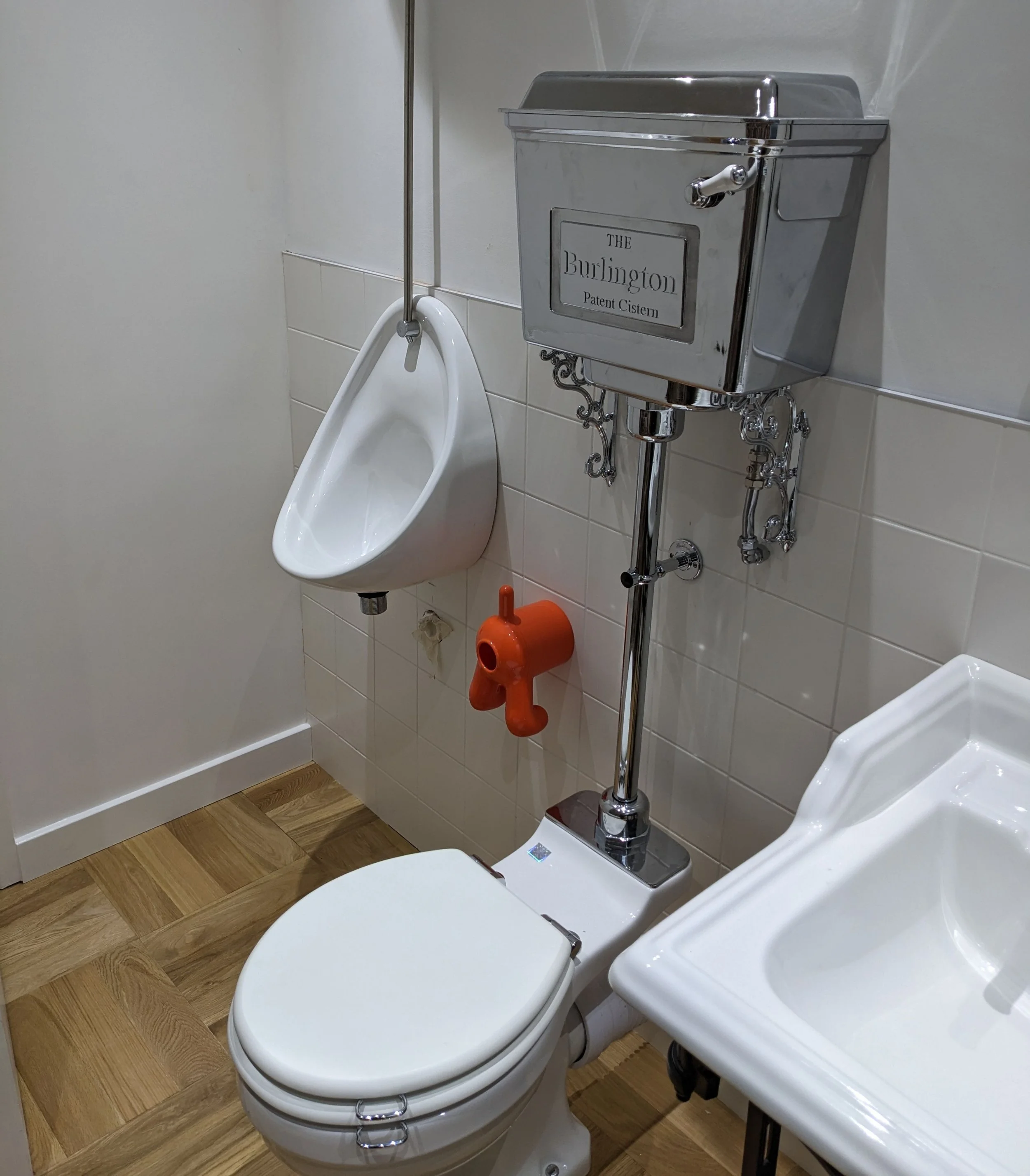 Qube Bathrooms. A small bathroom with a urinal, a toilet, and a sink. The urinal is mounted on the wall, and the toilet has a metallic cistern labeled 'The Burlington Patent Cistern'. The sink is partially visible in the lower right corner.