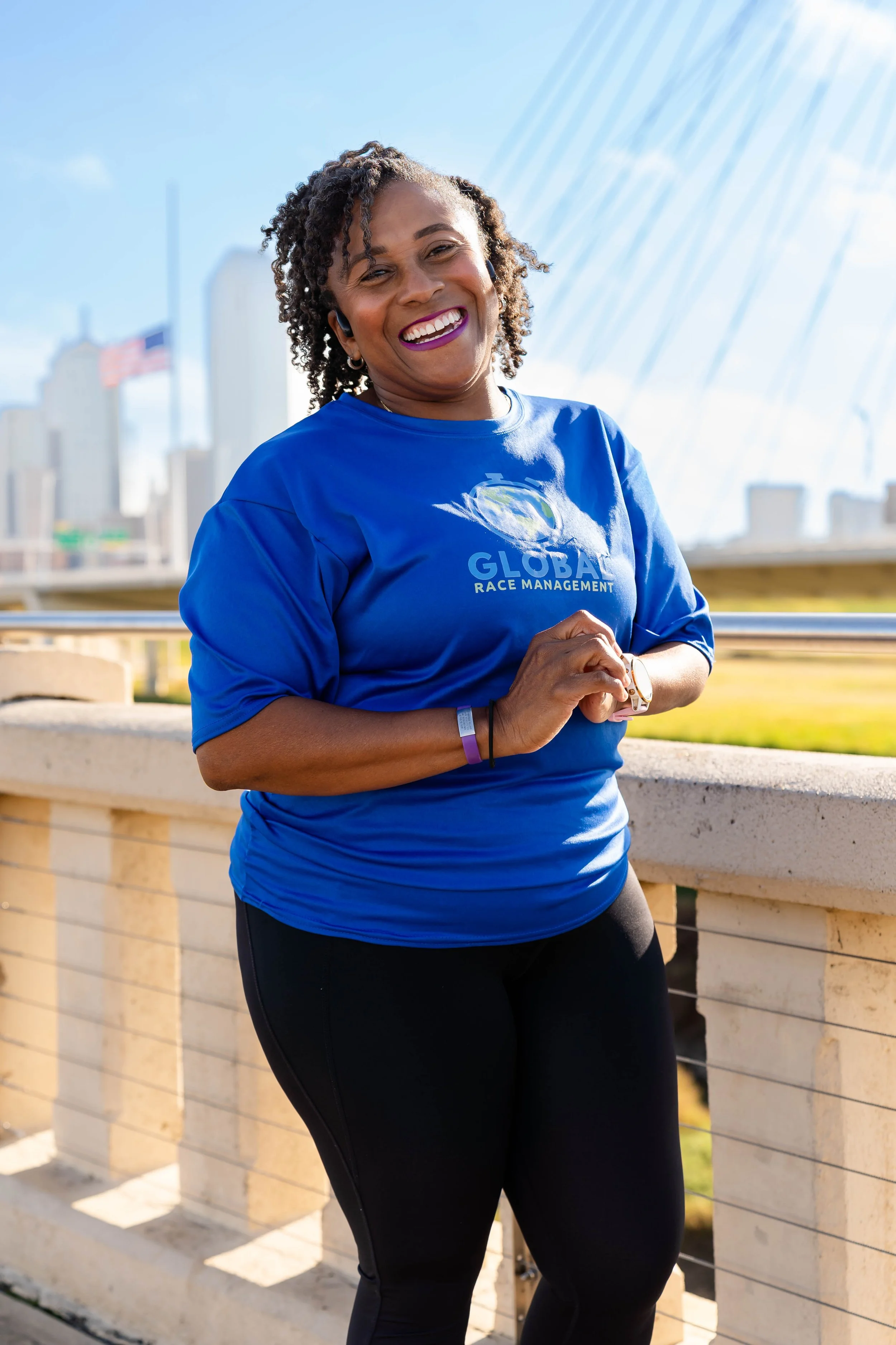 A woman smiling outdoors in running gear, wearing a blue t-shirt with a logo that reads 'GLOBAL RACE MANAGEMENT', sunglasses on her head, and earbuds, with a city skyline and a bridge in the background.