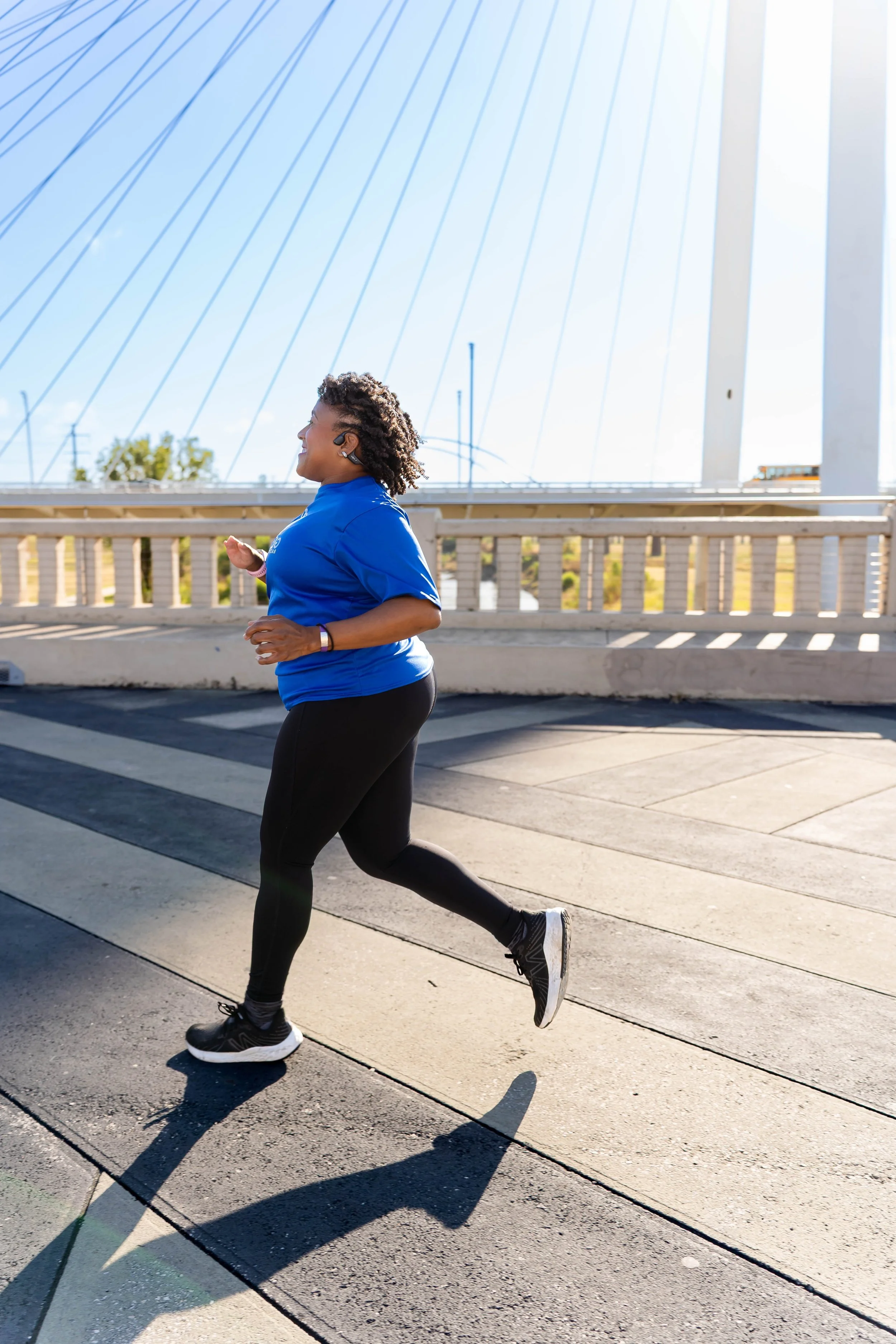 A woman jogging outdoors on a sunny day, wearing a blue shirt, black leggings, and black running shoes.