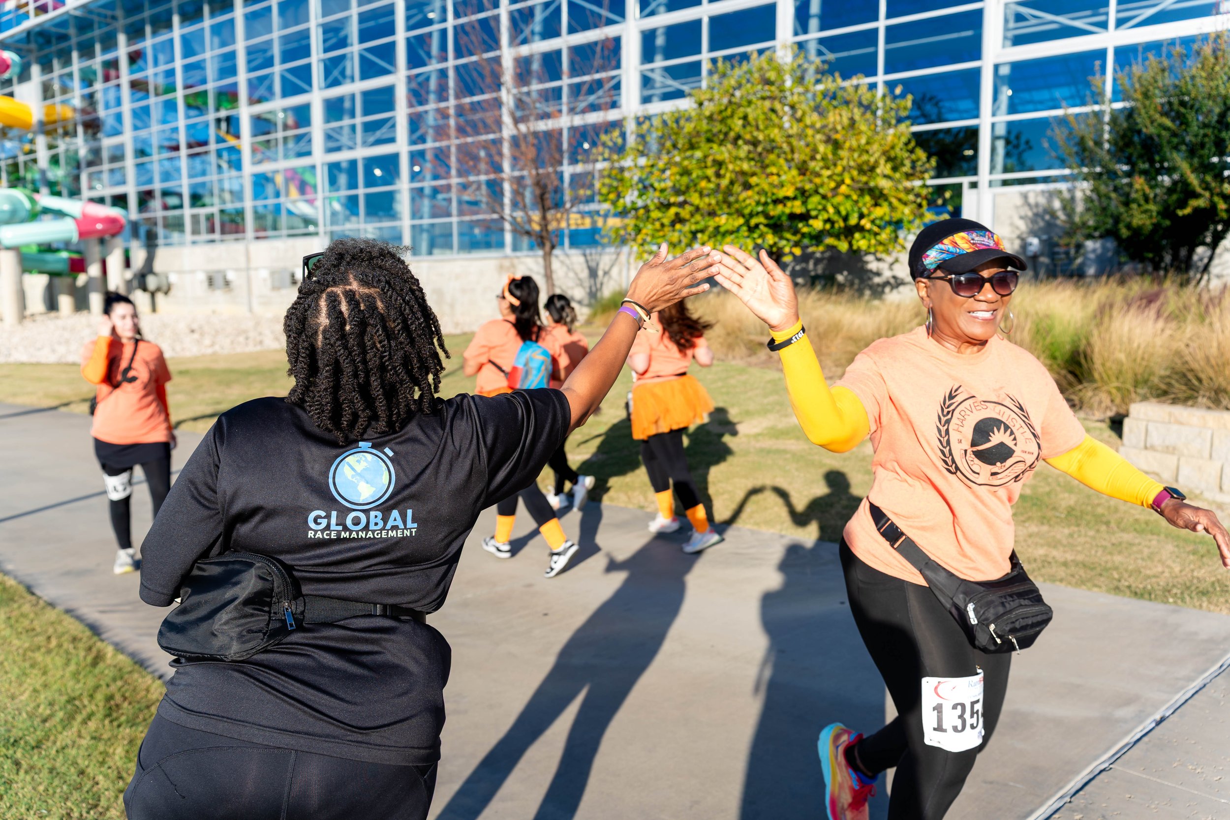 A woman high-fives a volunteer or supporter during a running event, with children running in the background, outside a modern building with colorful playground equipment.