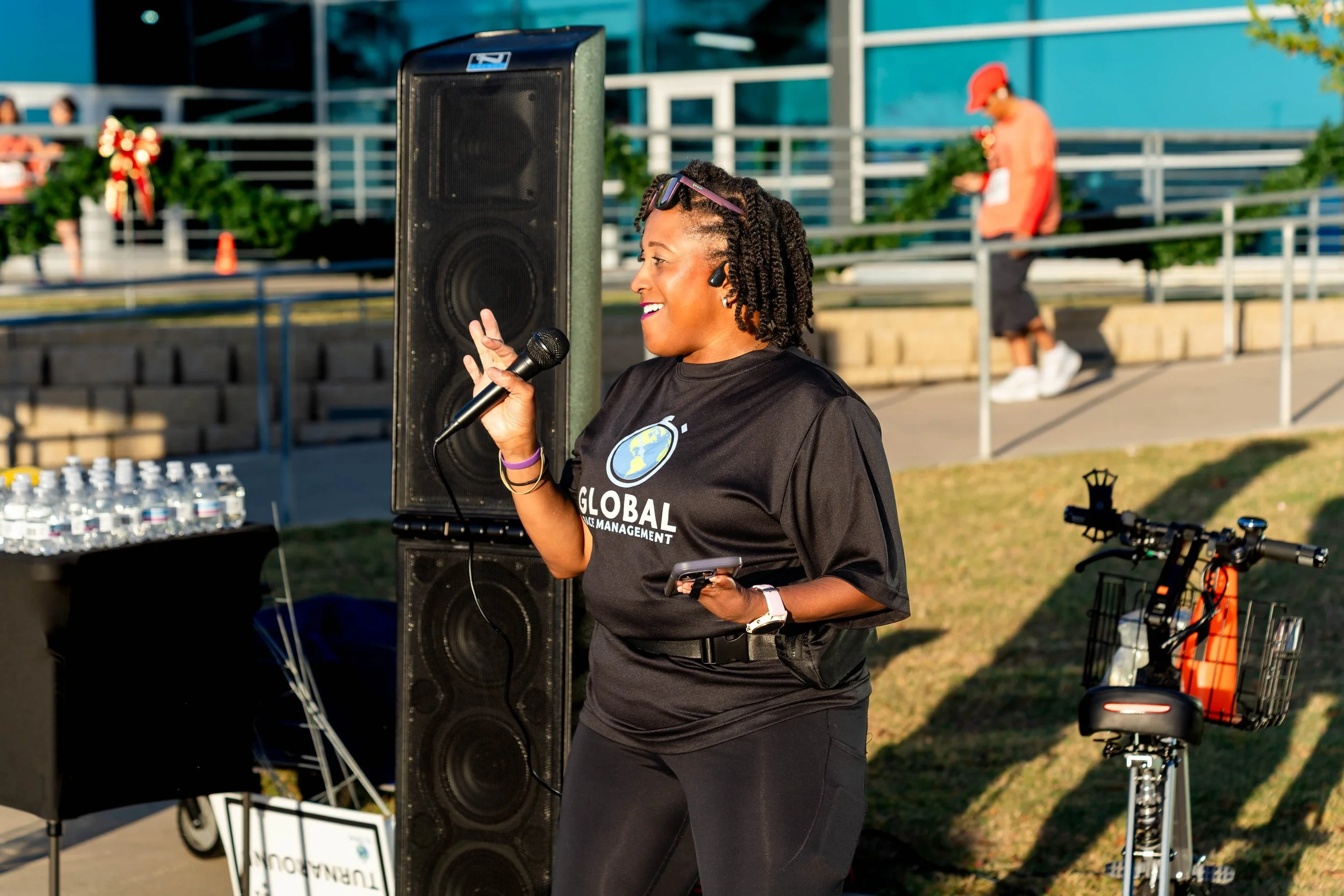 A woman speaking into a microphone at an outdoor event, wearing a black T-shirt with 'Global' written on it, standing next to a large speaker, with water bottles on a table nearby, and a bicycle with a basket in the background.