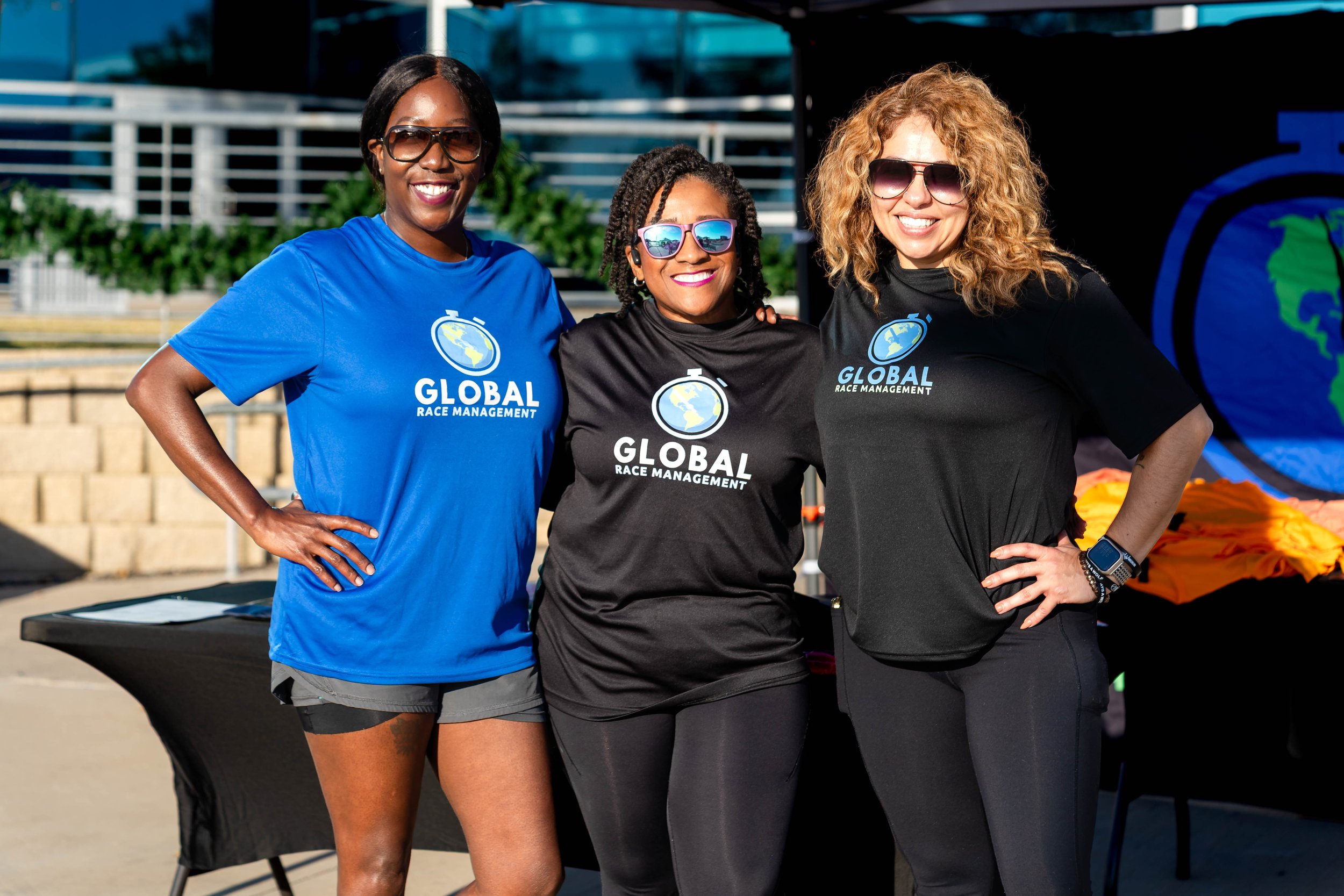 Three women smiling and posing for a photo at an outdoor event, wearing matching black and blue T-shirts that say 'Global Race Management.'
