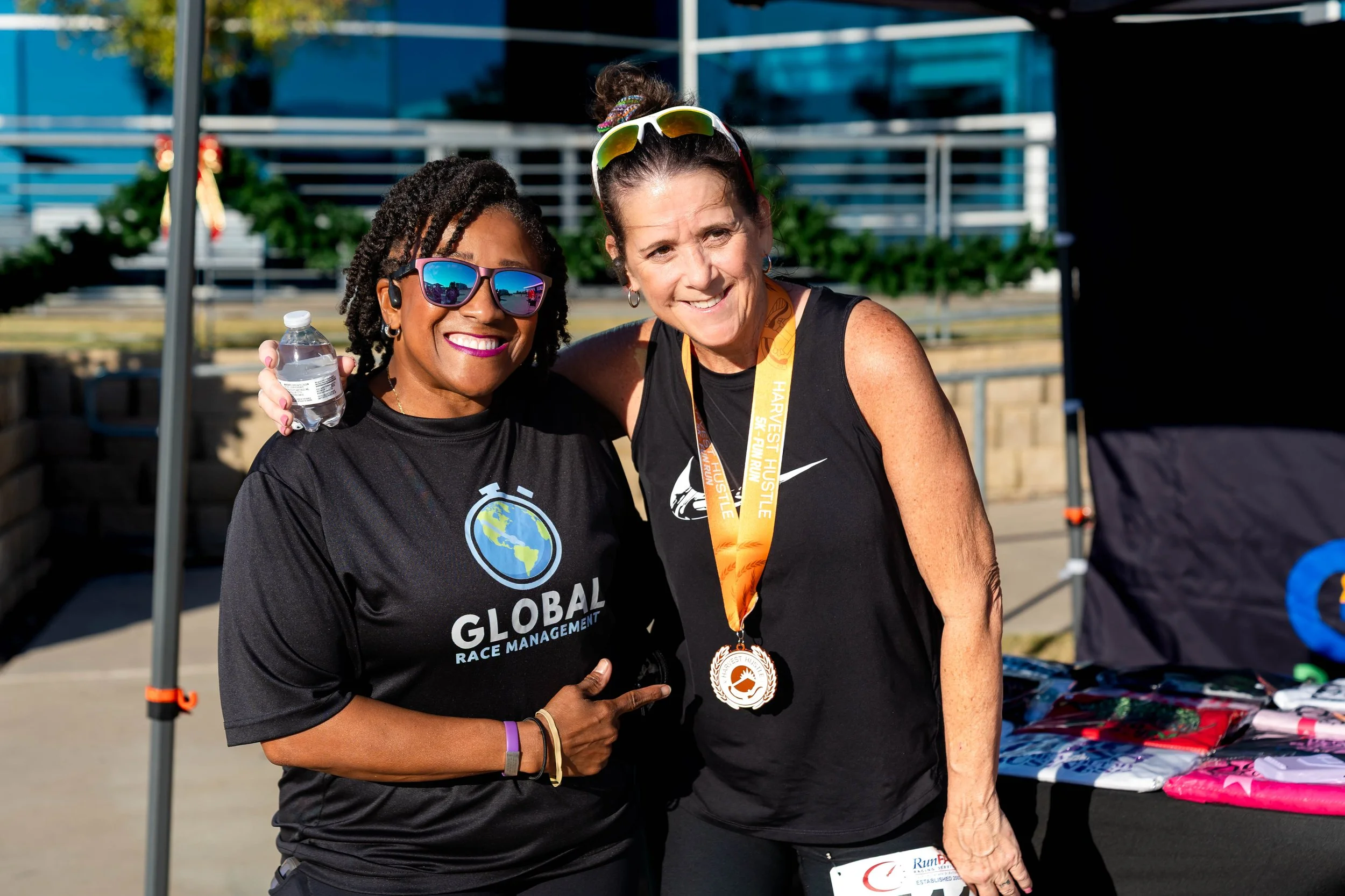 Two women smiling at a race event, one holding a water bottle and the other wearing a finisher medal around her neck, standing outdoors near a table with race merchandise.