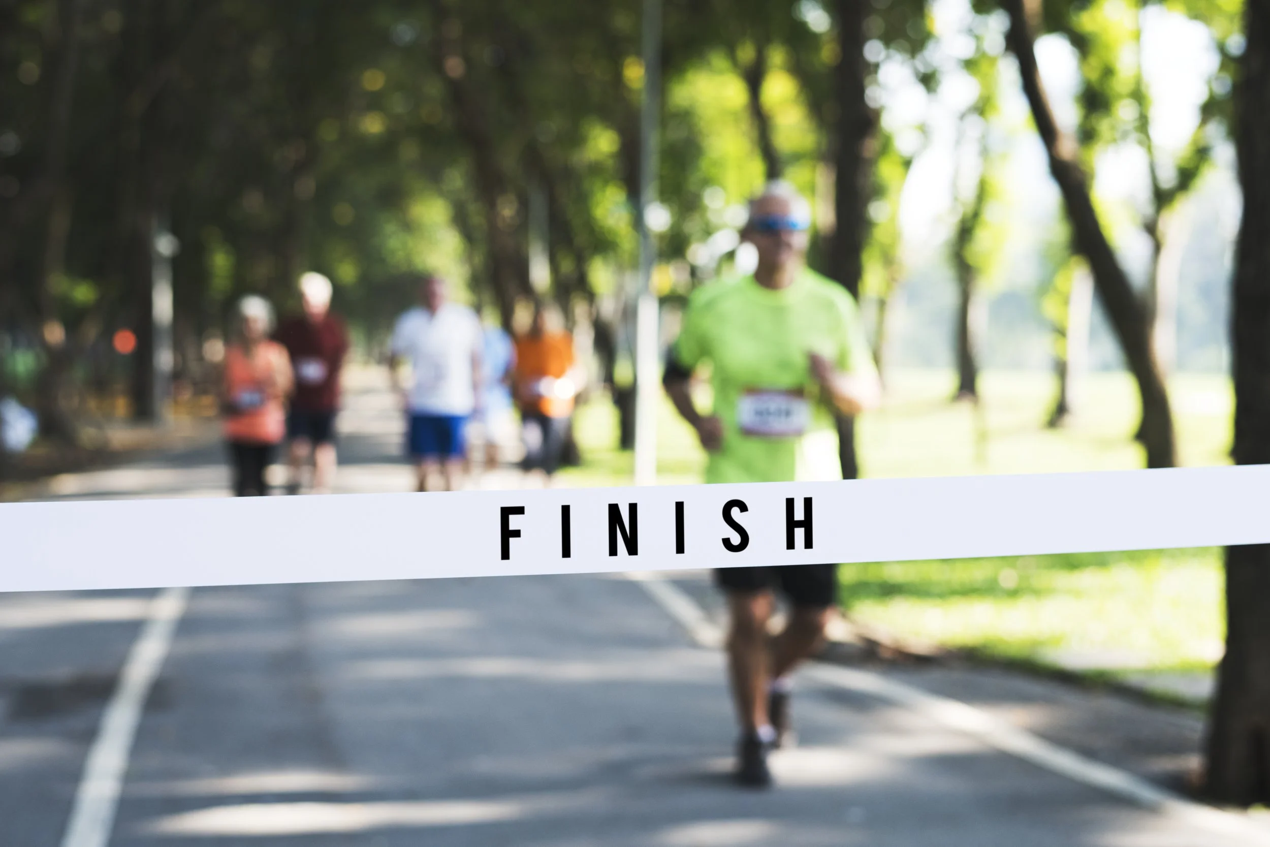 Runners finishing a race crossing the finish line in a park with trees.