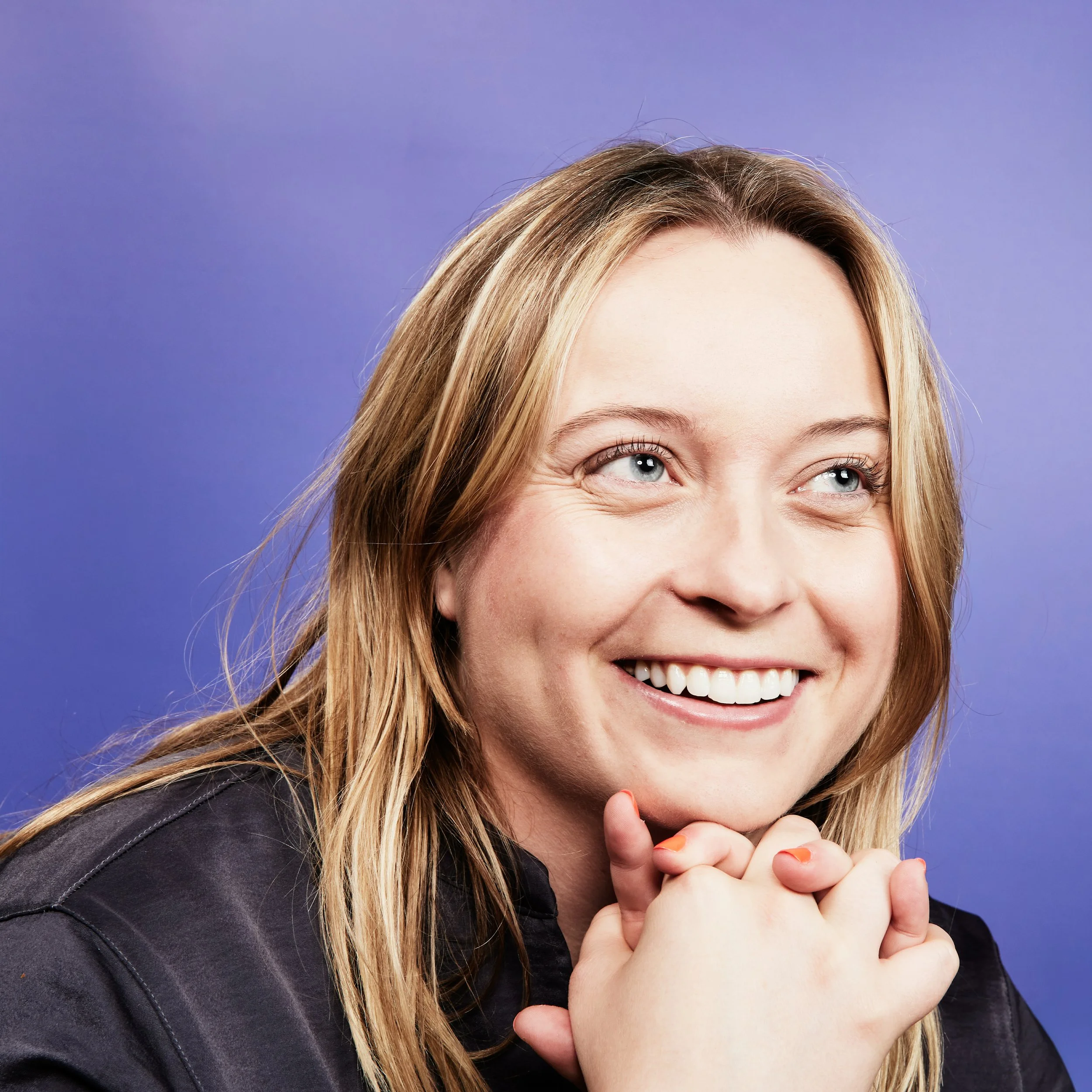 Smiling woman with blonde hair and blue eyes, wearing a navy shirt, against a purple background.