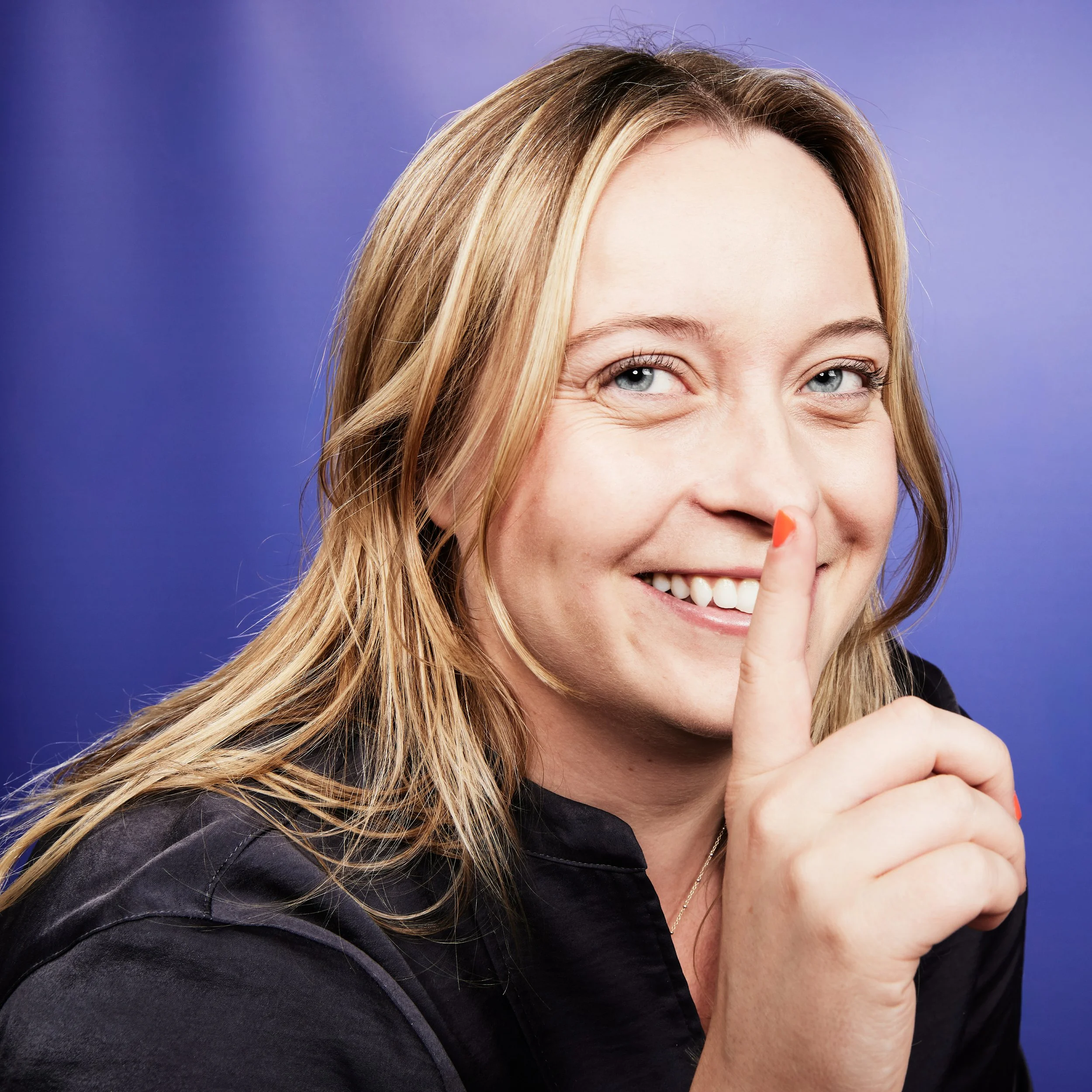 A woman with blonde hair and blue eyes smiling, holding her index finger up in the air against a purple background.