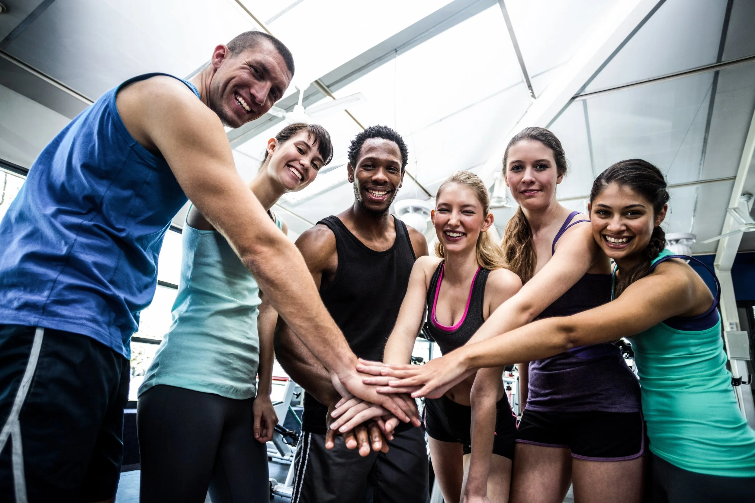 Group of six diverse young adults in athletic wear smiling and placing their hands together in a gym.