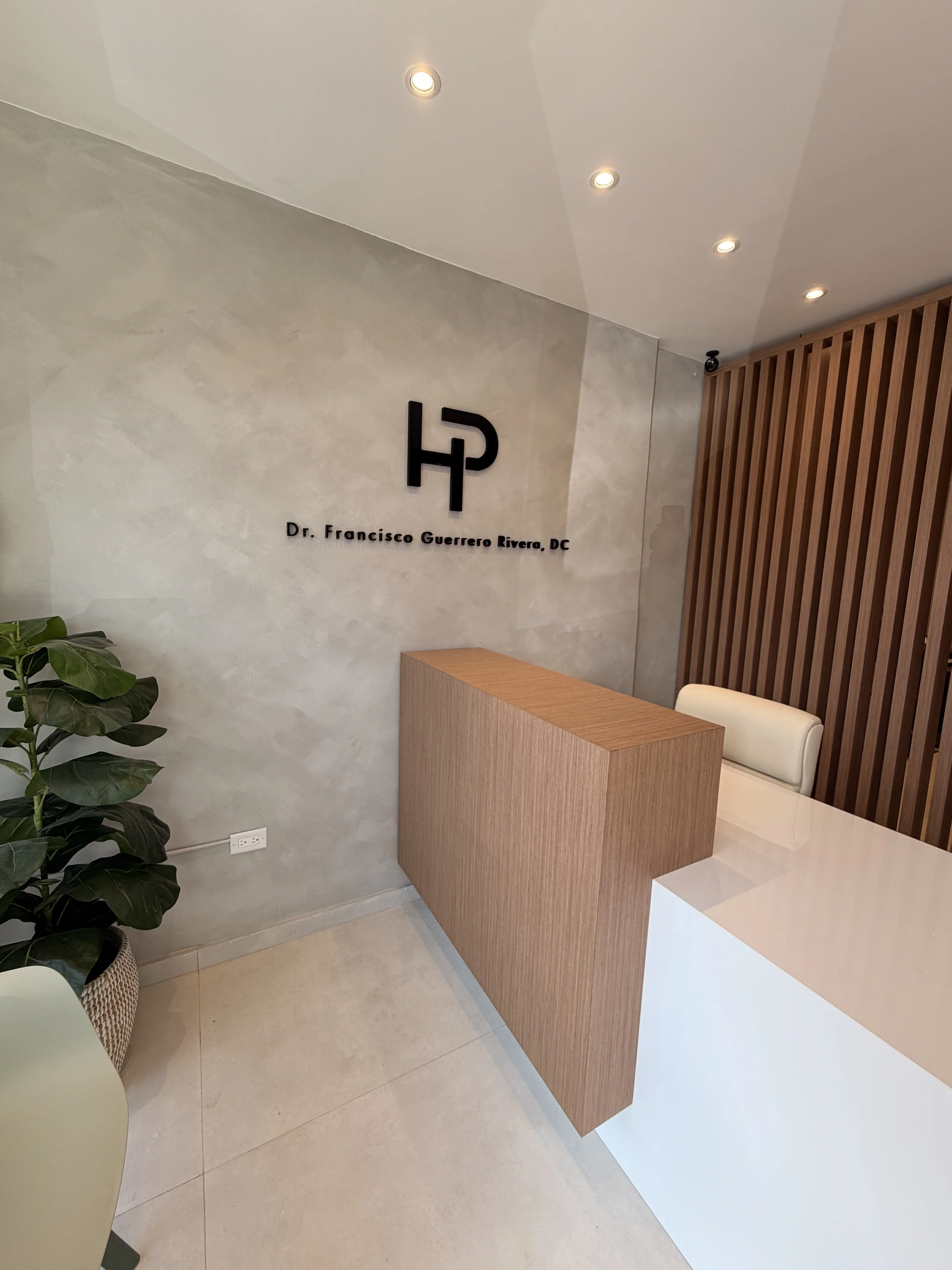Reception area with a wooden desk, a white chair, a green potted plant, and a decorative wall with a logo and the name Dr. Francisco Guerrero Rivera, DC.