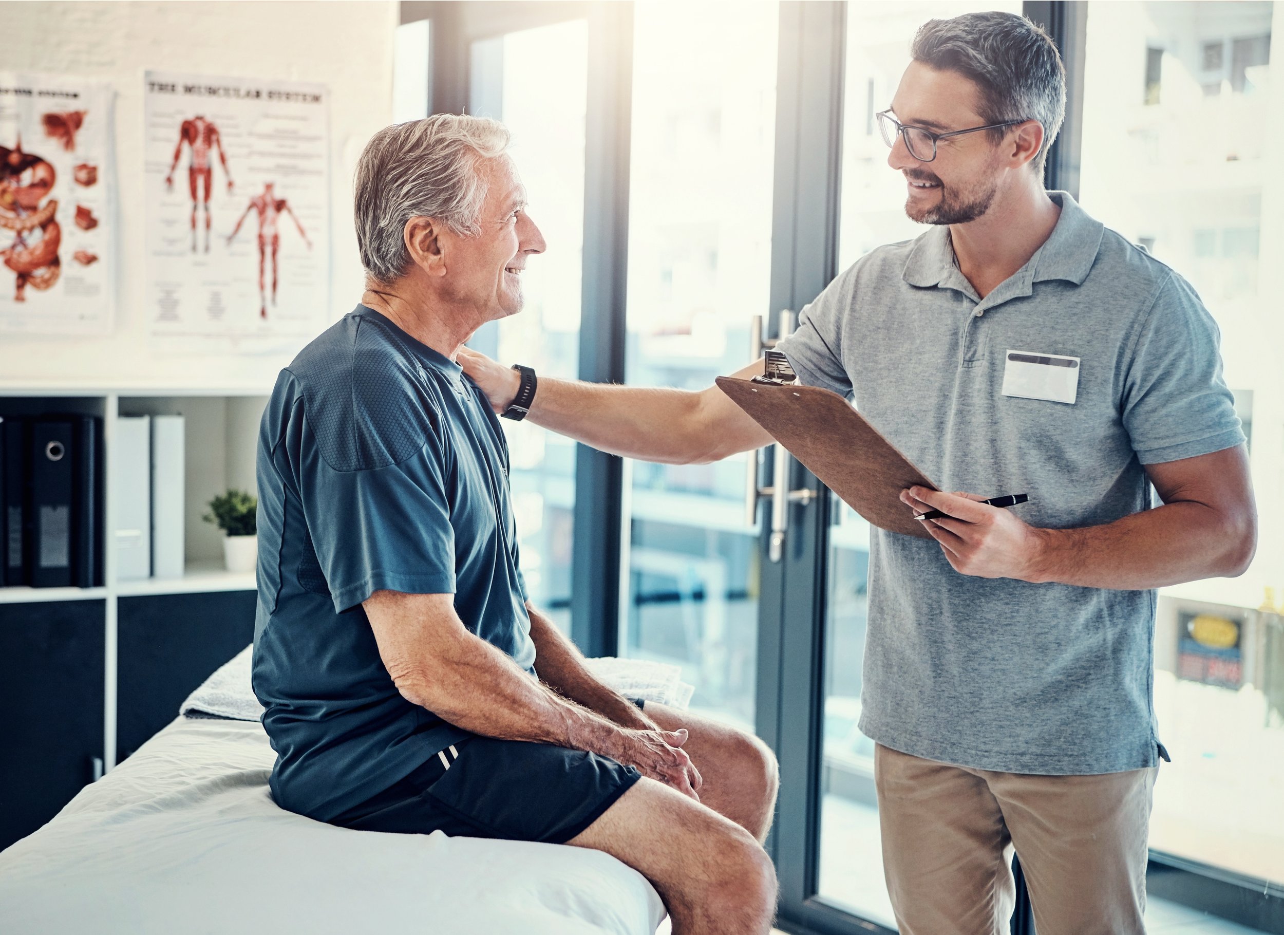 A doctor or healthcare professional examining an elderly man who is sitting on a medical bed. The professional is smiling and holding a clipboard, while the elderly man, dressed in athletic wear, is also smiling. The setting appears to be a medical or physical therapy office with anatomical posters on the wall.