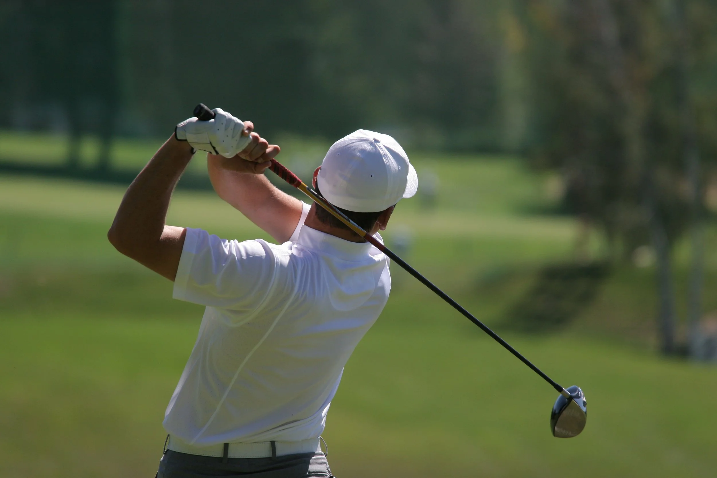 Male golfer in a white shirt and cap swings a golf club on a golf course with green grass and trees in the background.