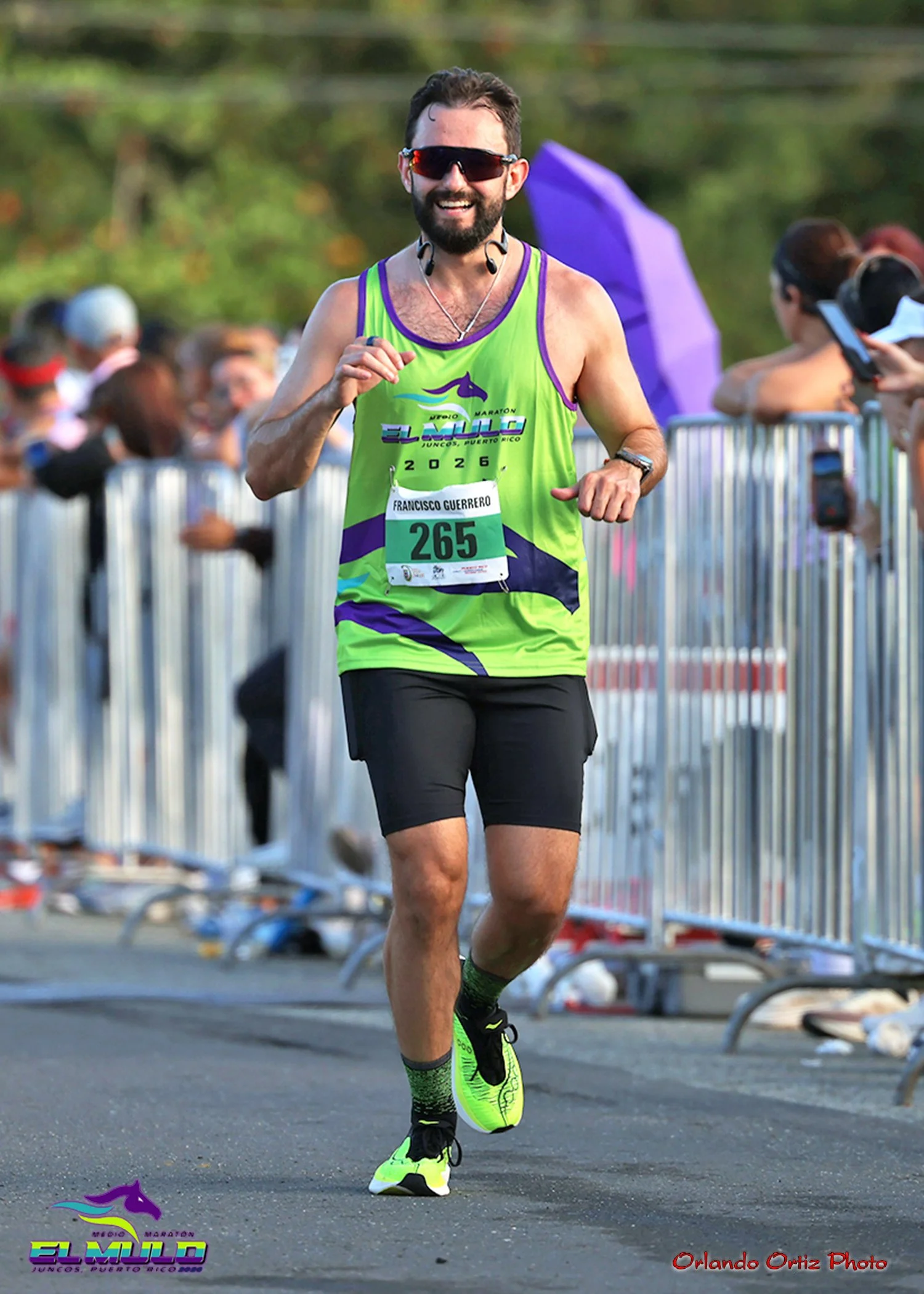 A man running in a marathon, smiling, wearing sunglasses, a green and purple running tank top, black shorts, neon yellow running shoes, and a racing bib numbered 265.