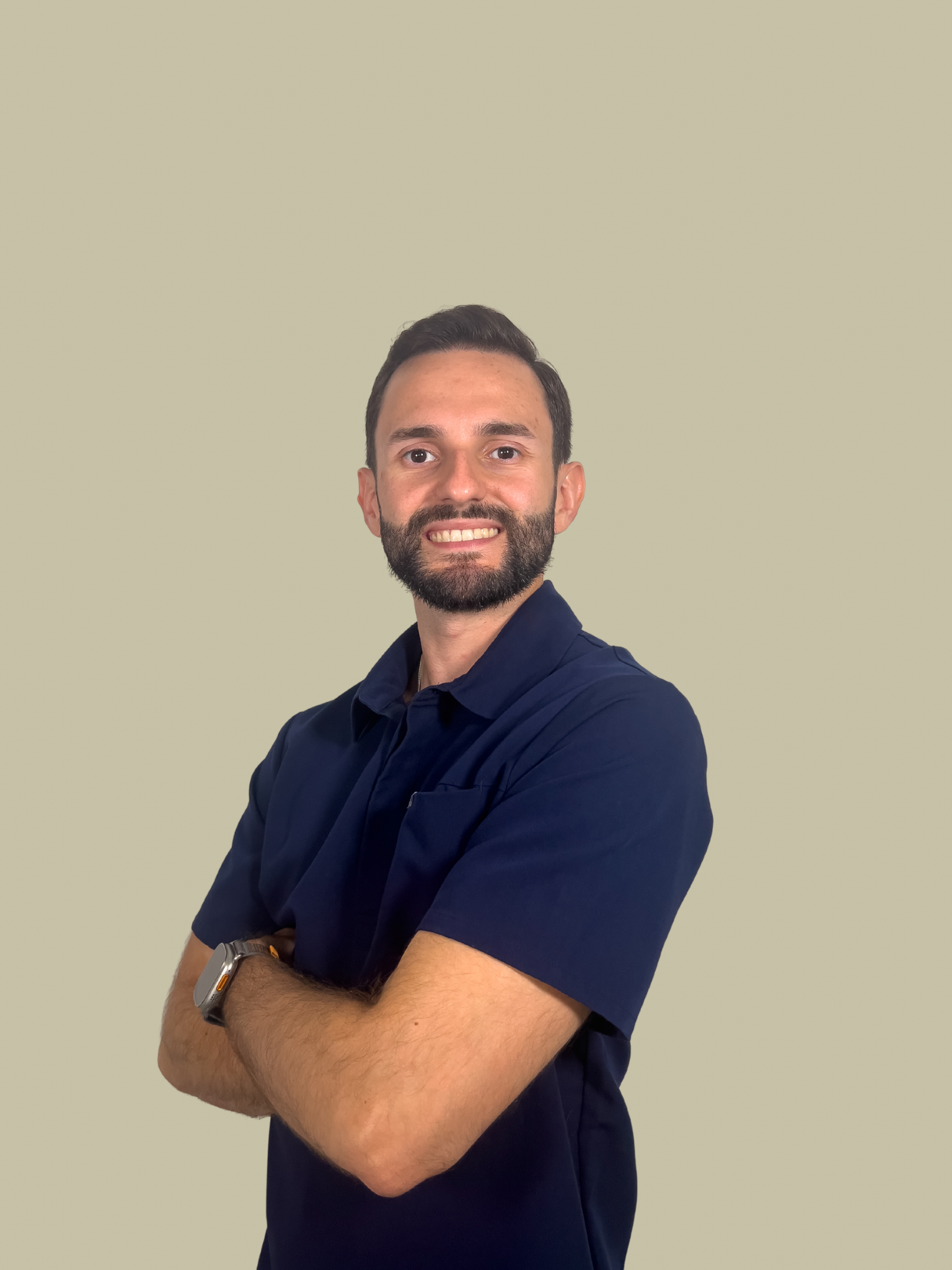 Dr. Guerrero is smiling with a beard, wearing a navy blue short-sleeve shirt, standing with crossed arms against a plain light-colored wall.