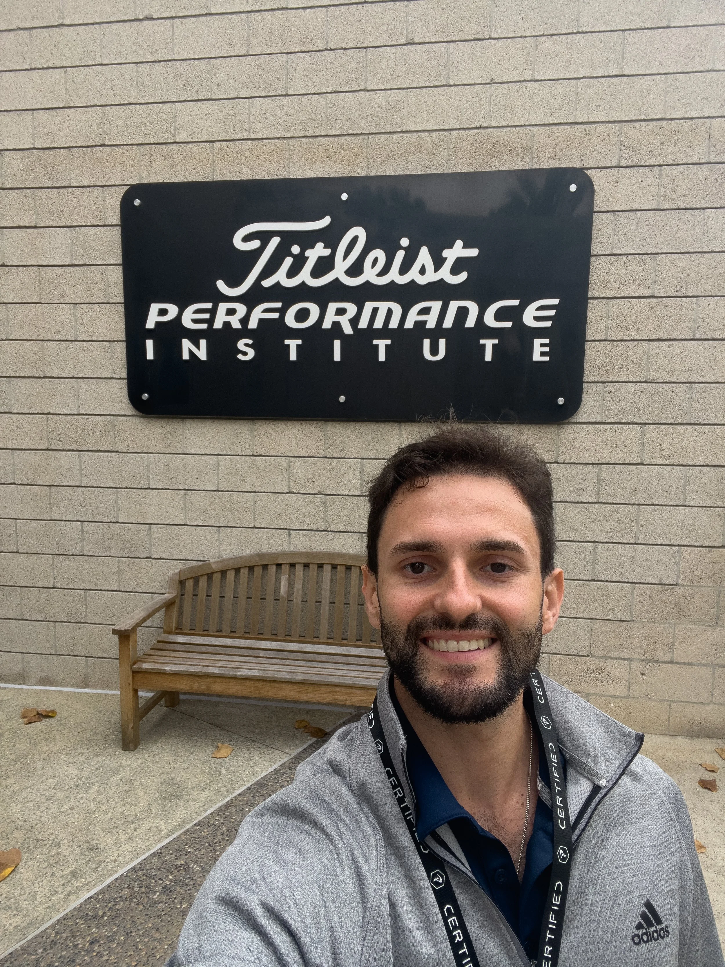 Dr. Guerrero taking a selfie in front of a sign that reads 'Titleist Performance Institute' on a brick wall, with a wooden bench nearby.