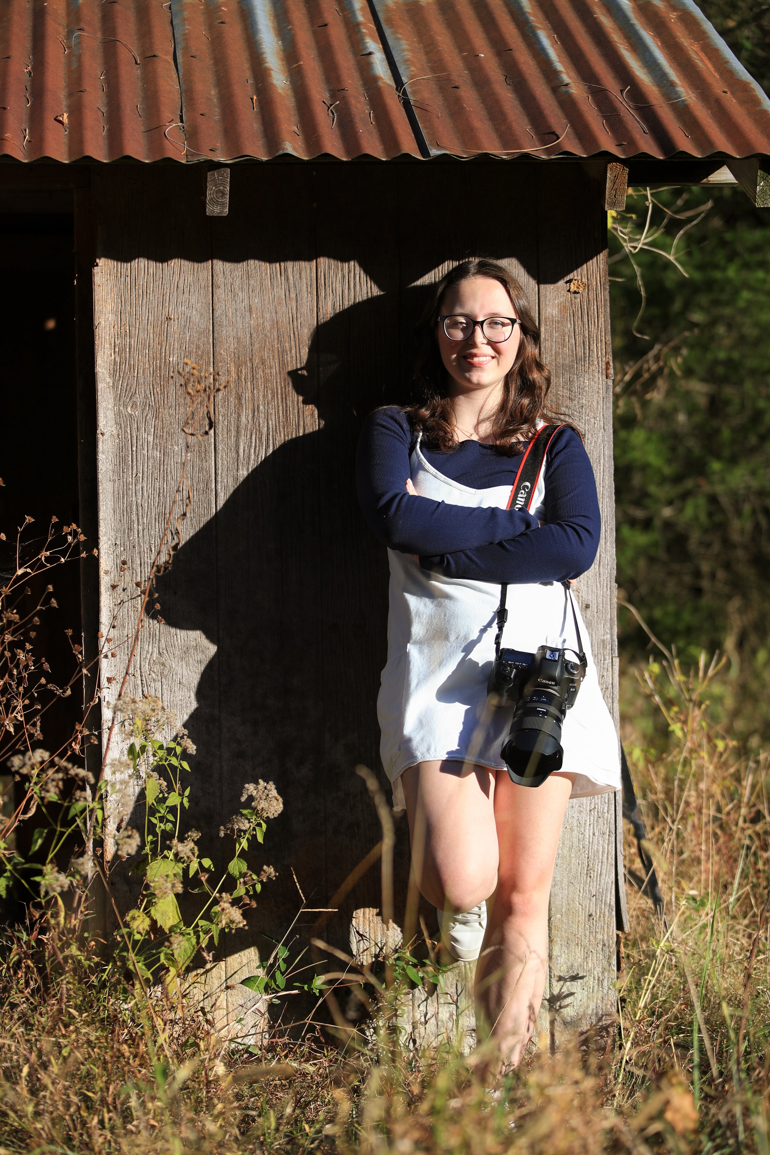 A woman with glasses and dark hair smiling, standing outdoors against a rustic wooden wall, with a camera hanging from her neck, surrounded by tall grass and plants.
