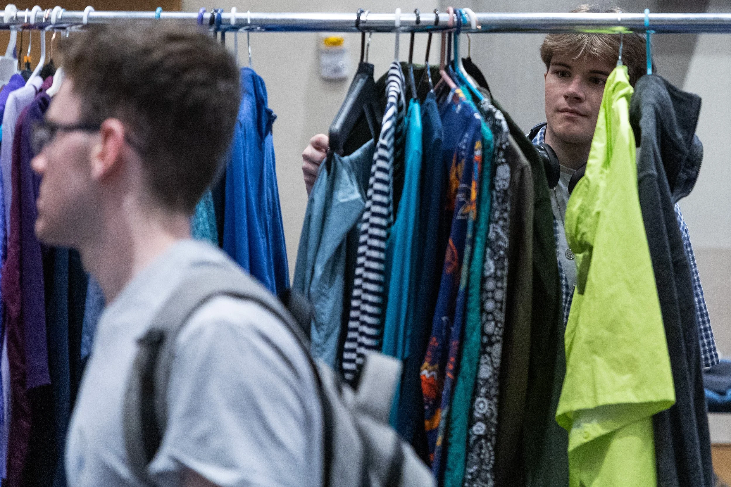 Two young men shopping through a rack of colorful clothing, with one in the foreground wearing glasses and a backpack, and the other in the background looking at the clothes.