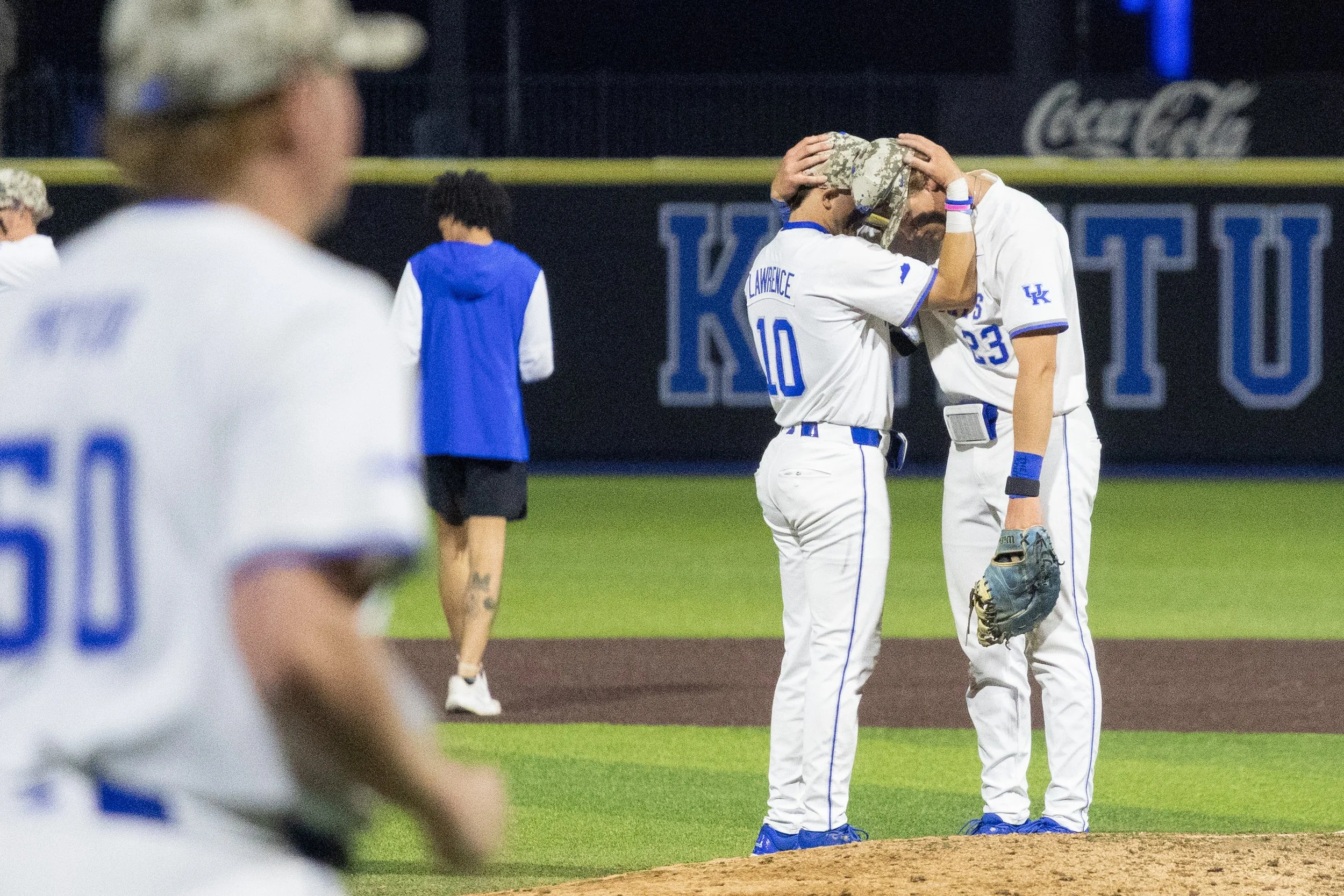 Two baseball players in white uniforms, one with the number 10 and the other with 23, are standing close together on the pitcher's mound, with one player holding his head in his hands. Two other players are visible in the background, one with only partially visible uniform. The scene is lit up as if during a game night.