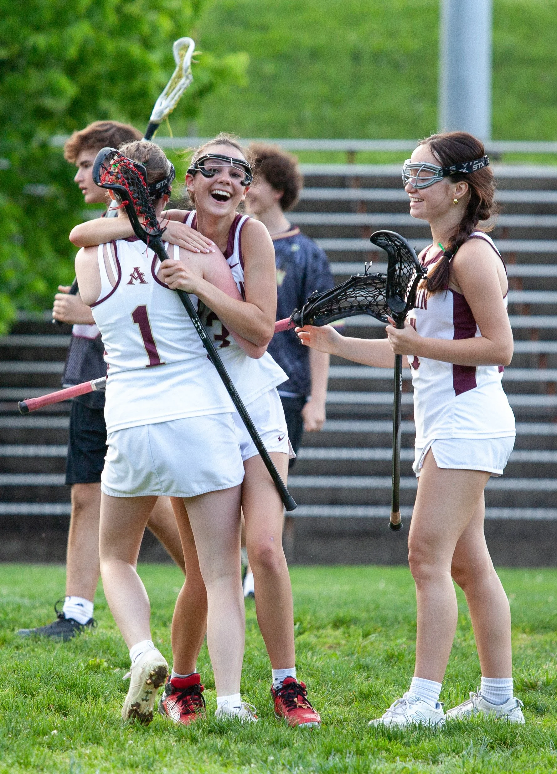 Lacrosse players in white uniforms celebrating on the field, with one player hugging another while holding lacrosse sticks, and two other players smiling nearby.