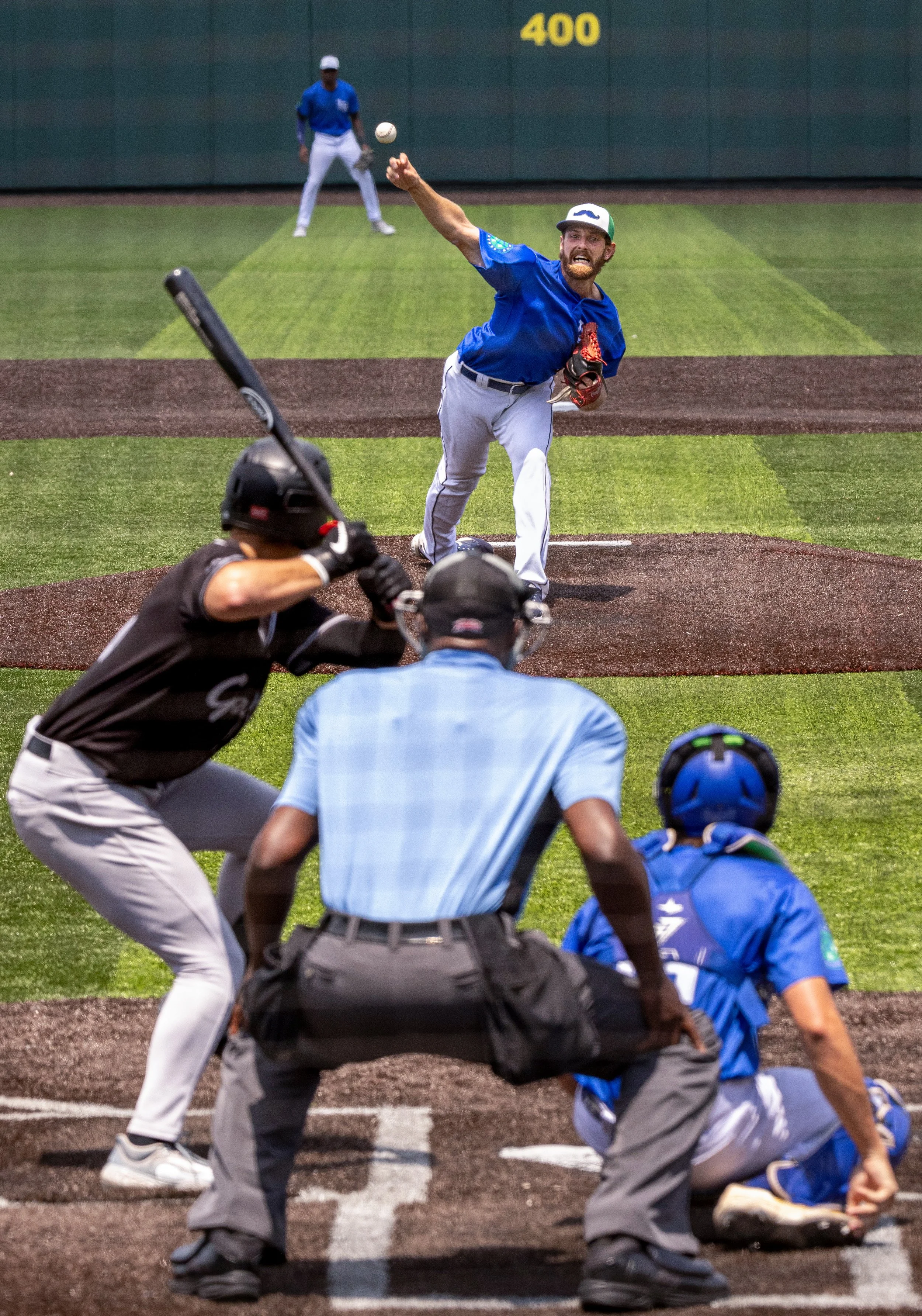 A baseball player in a blue uniform throws a pitch from the mound at a baseball field, with the home plate umpire, catcher, and batter in position, and an outfielder in the background.