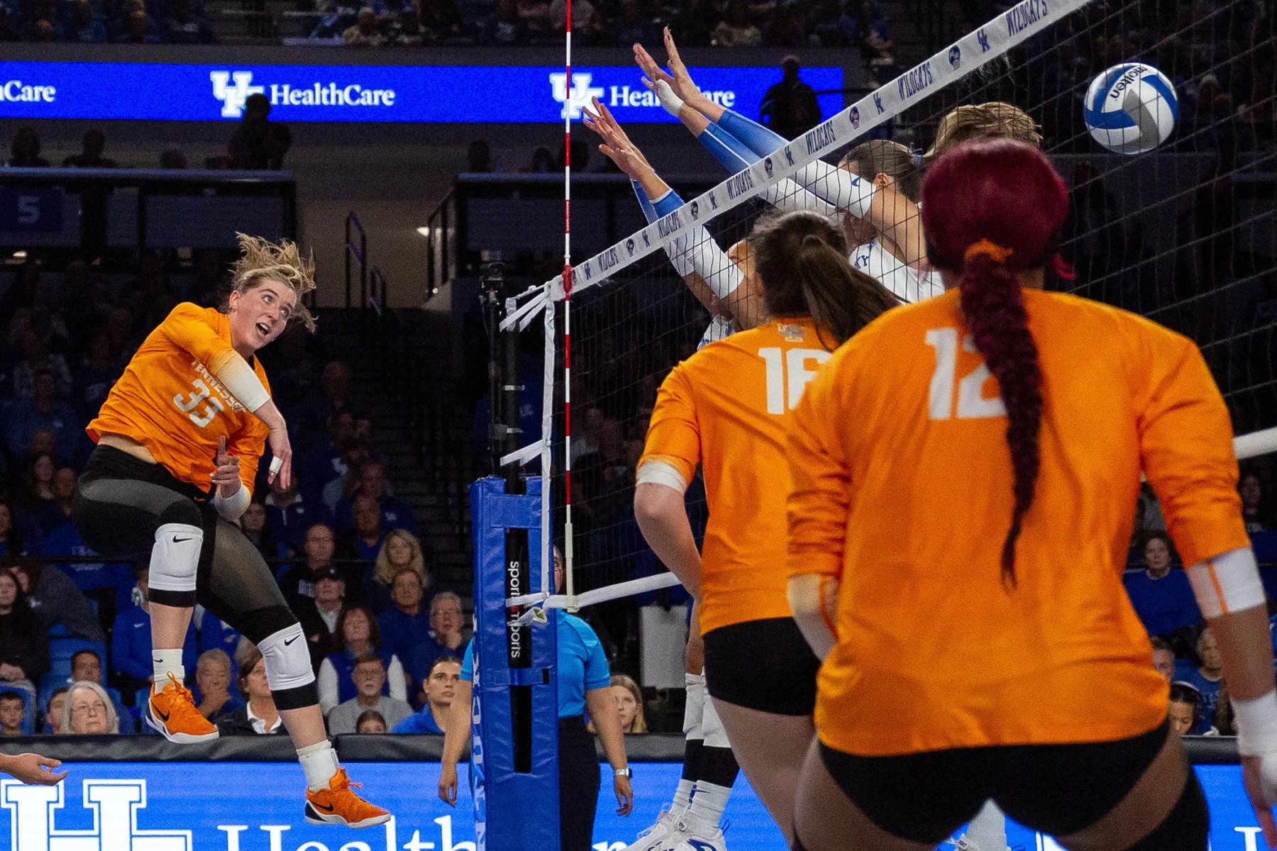 A volleyball game with players in orange jerseys blocking, and a player in an orange jersey jumping to hit the ball.