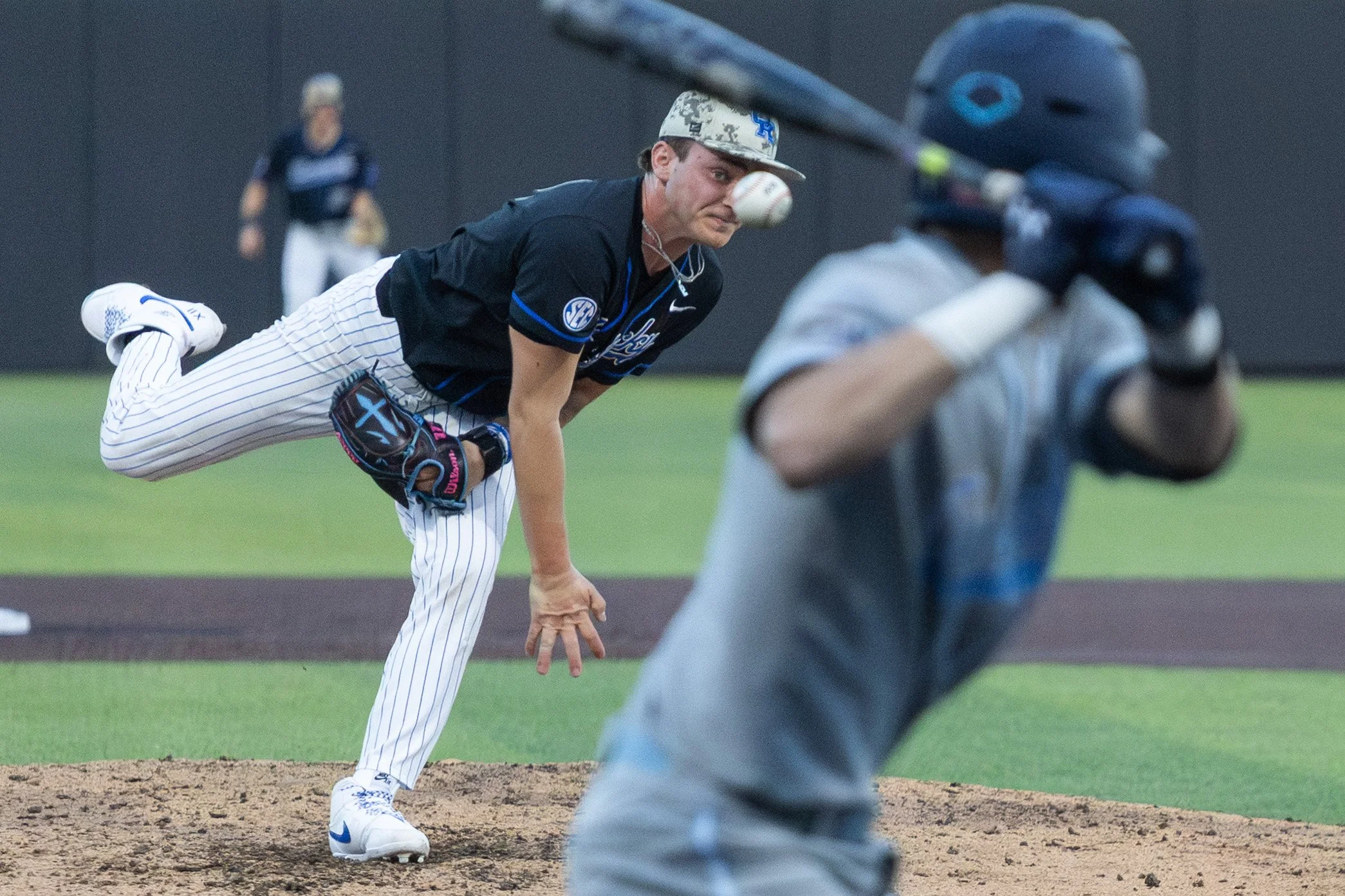 A baseball player in a black jersey and white pinstriped pants is pitching the ball from the mound, with a batter in a gray uniform ready to hit in the foreground.