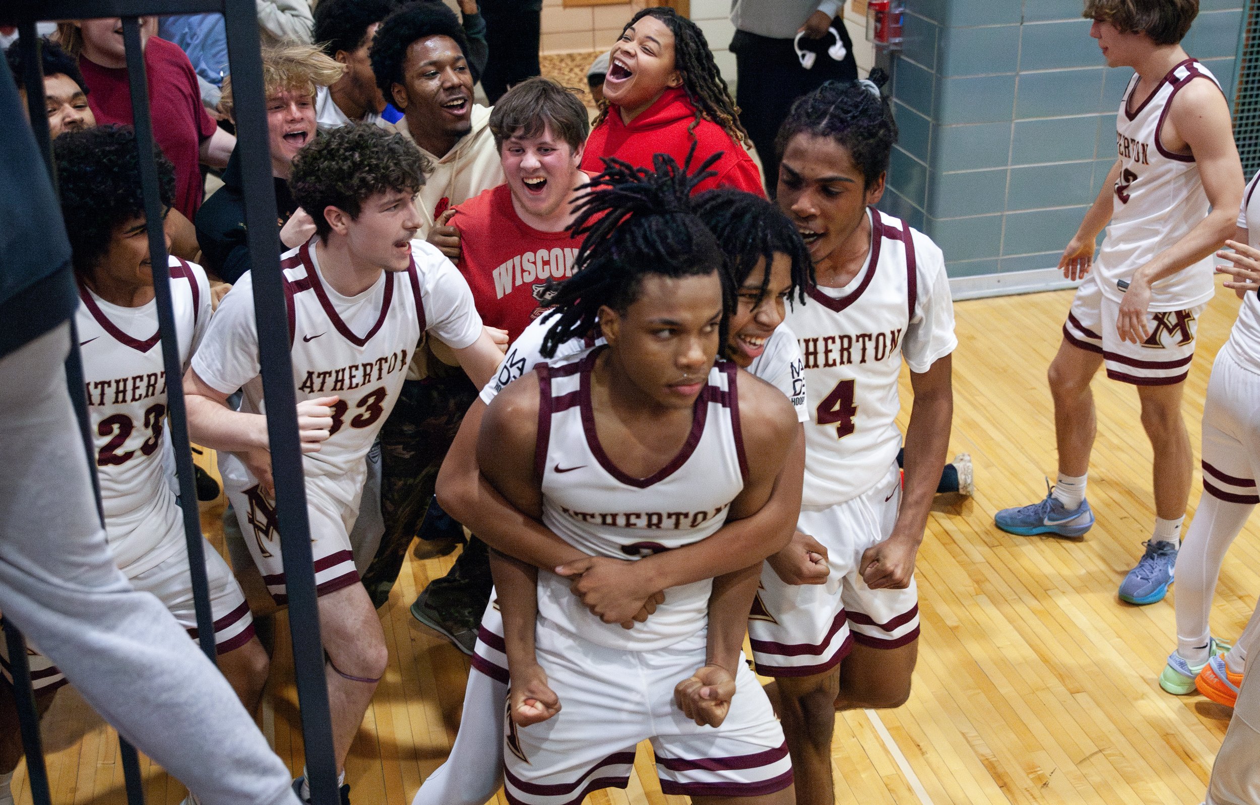 A group of basketball players in white jerseys with maroon trim, celebrating and cheering in a gymnasium. One player is in the foreground with a determined expression, while others in the background are smiling and shouting excitedly.