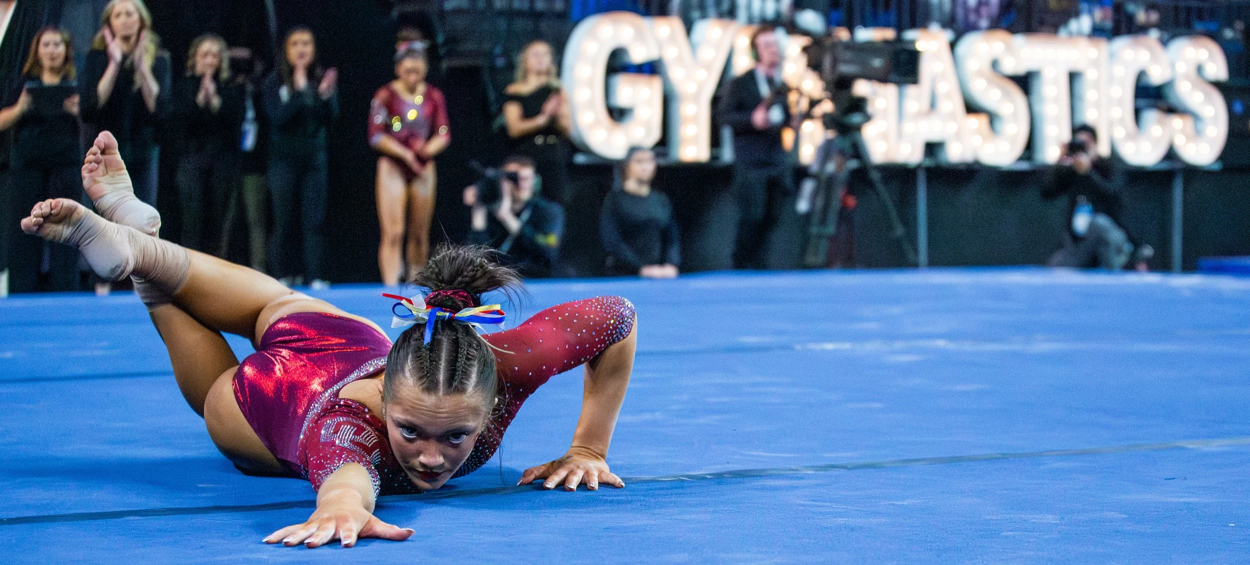 Young female gymnast in a red leotard performing a routine on a blue mat during a gymnastics competition, with an audience and large illuminated 'GYMNASTICS' sign in the background.