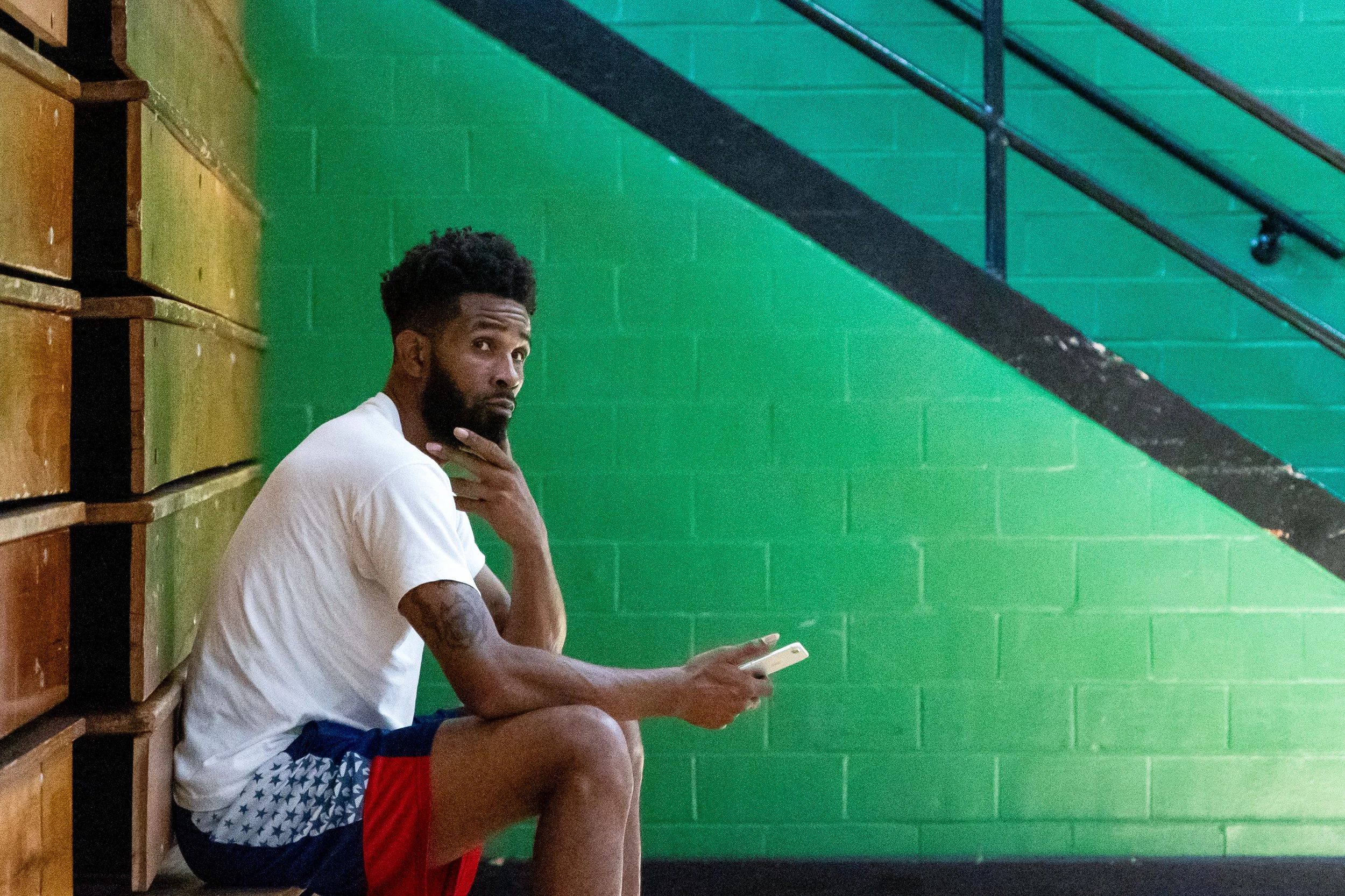 A man with dark hair and beard, wearing a white t-shirt and athletic shorts, sitting on wooden benches against a green brick wall, holding a phone in his right hand and resting his chin on his left hand, looking at the camera.