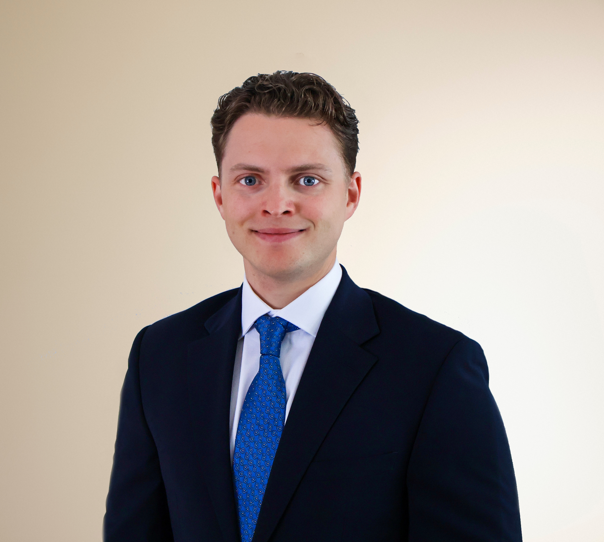 A young man with short, curly brown hair and blue eyes, wearing a dark navy suit, white dress shirt, and a blue patterned tie, standing against a plain light beige background.